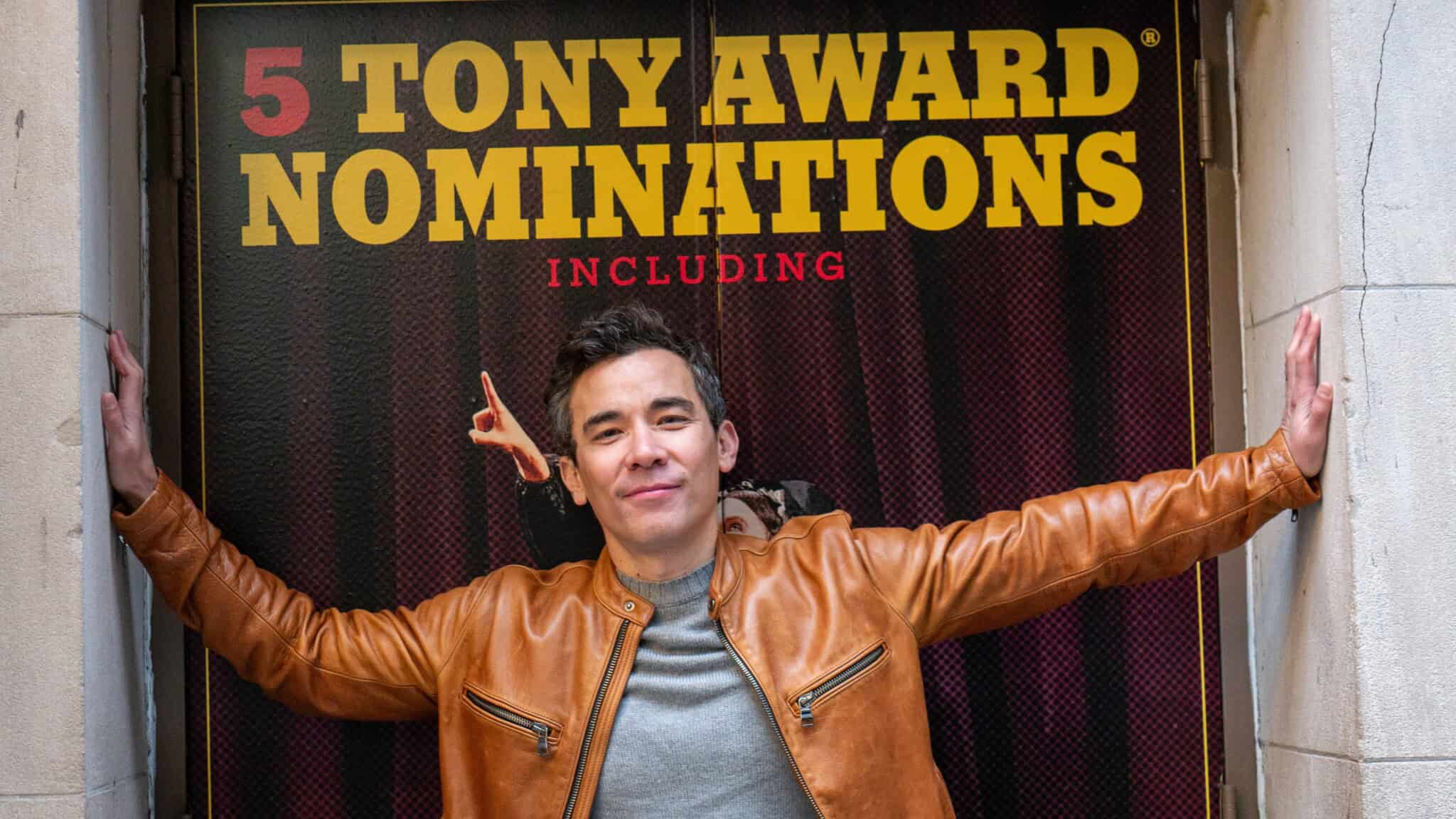 Conrad Ricamora stands below a sign that reads “5 Tony Award Nominations Including”