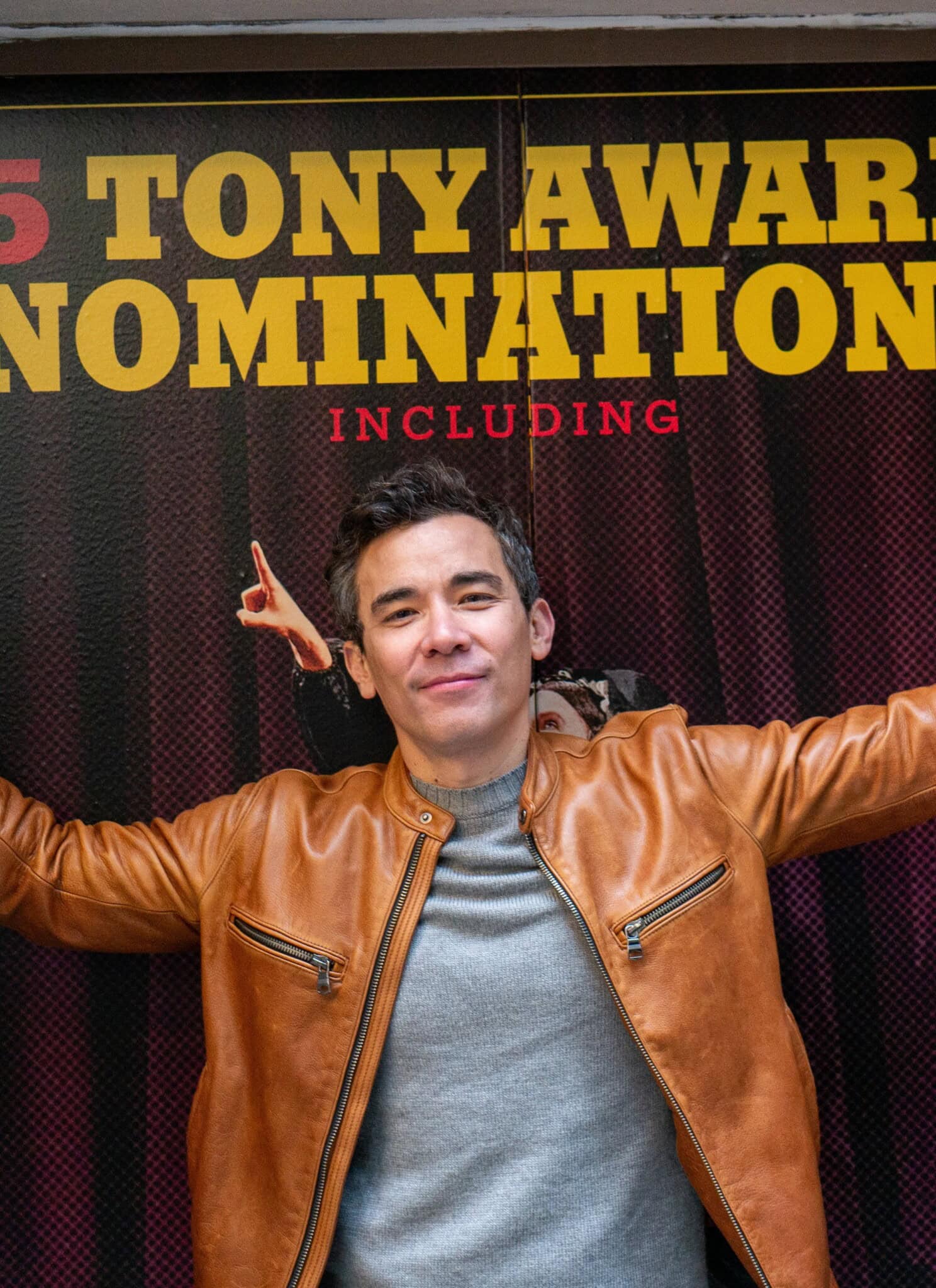 Conrad Ricamora stands below a sign that reads “5 Tony Award Nominations Including”