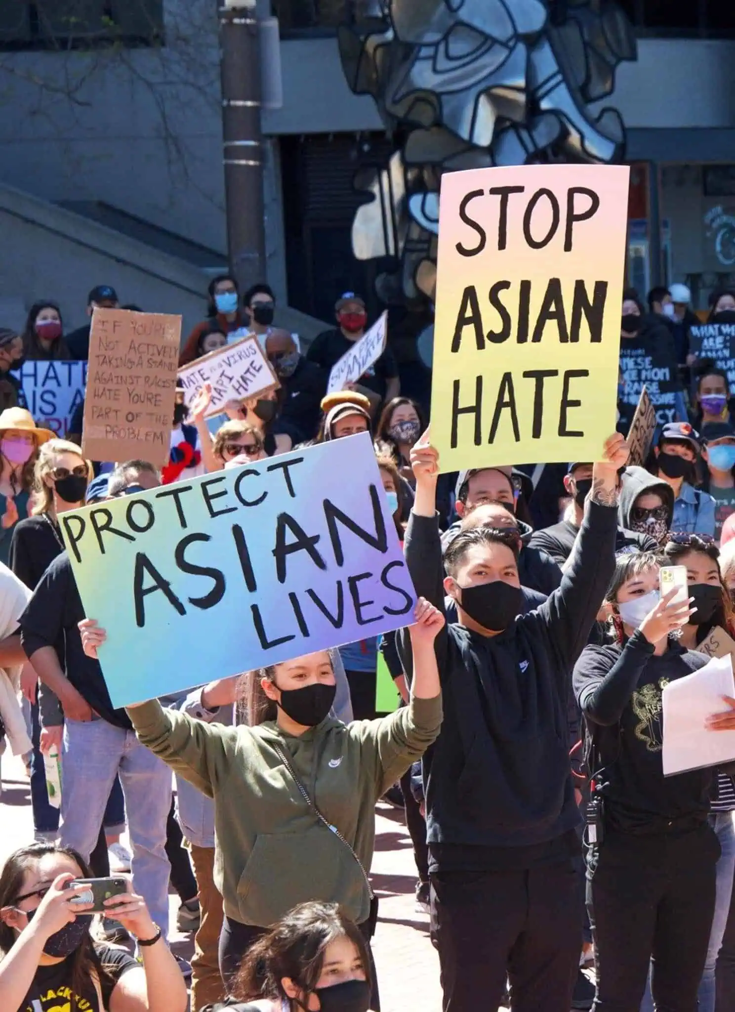 Predominantly Asian protesters in face masks stand together, holding up signs about anti Asian hate.