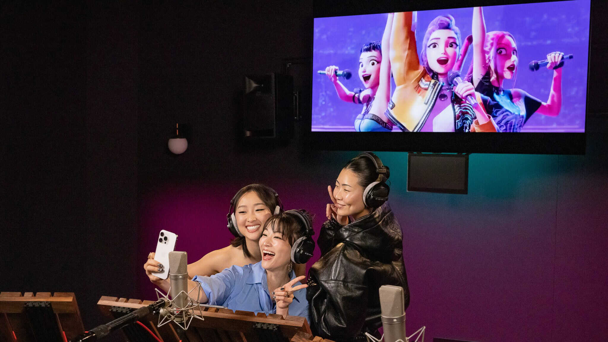 Three Asian women stand together in a recording booth, and pose for a selfie, with an animated film in the background.