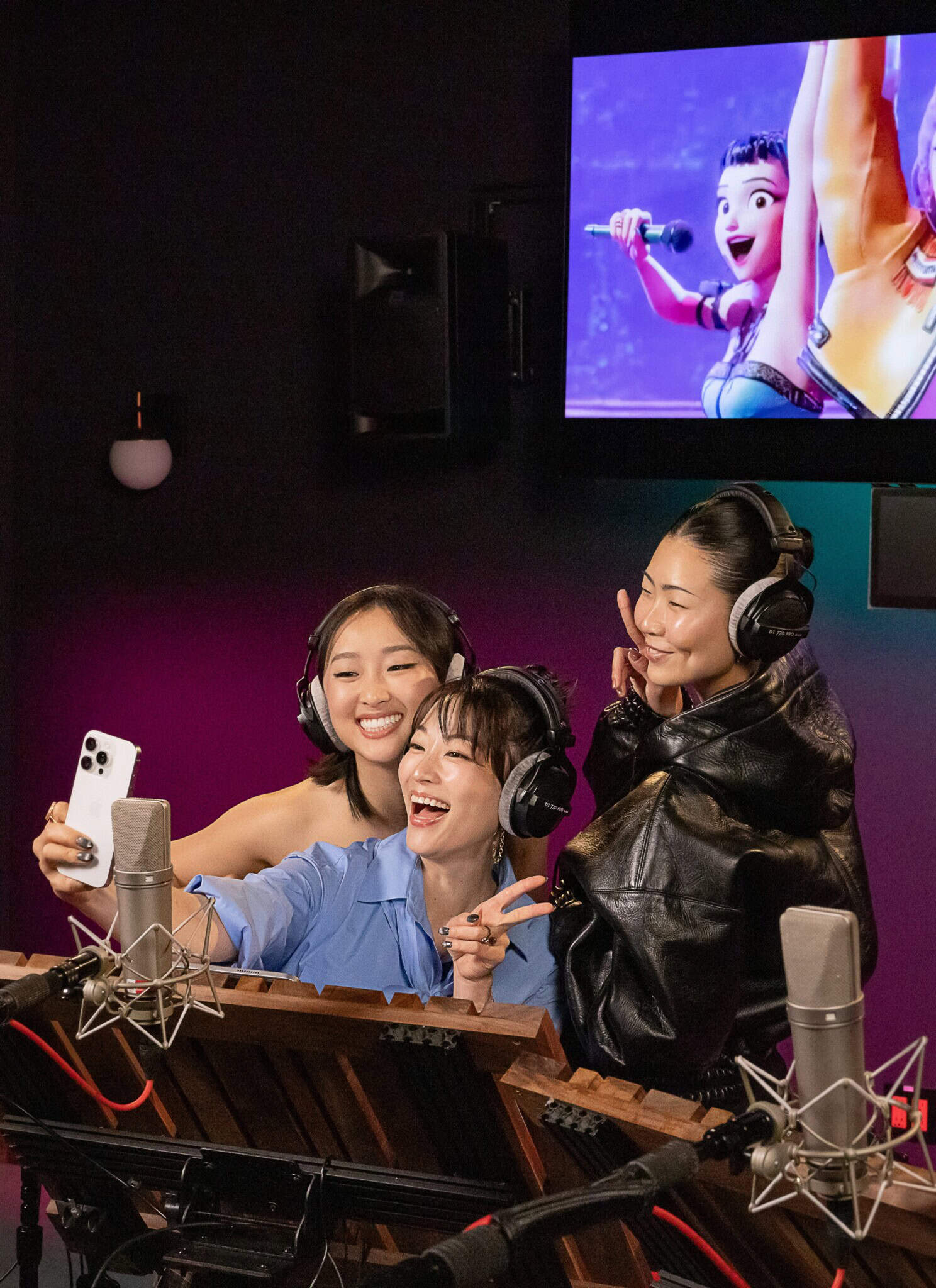 Three Asian women stand together in a recording booth, and pose for a selfie, with an animated film in the background.