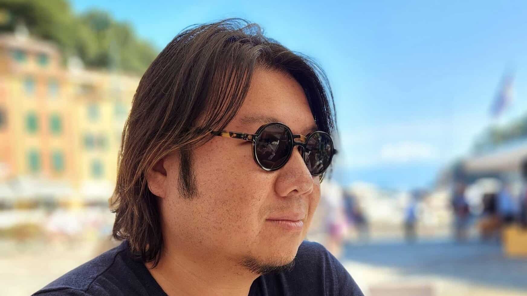 An Asian man in sunglasses and a navy blue shirt, with a blue sky and buildings in the background.