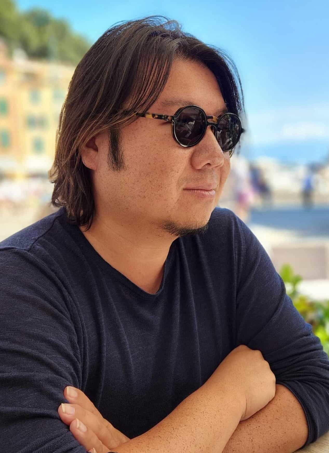 An Asian man in sunglasses and a navy blue shirt, with a blue sky and buildings in the background.