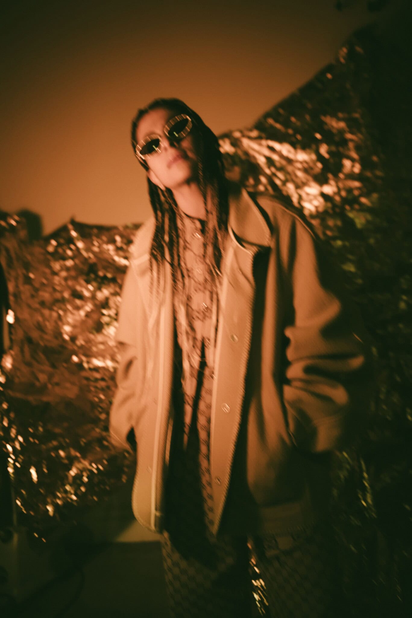 Musical artist Madame Gandhi, dressed in brown and beige, stands against a brow and gold backdrop.