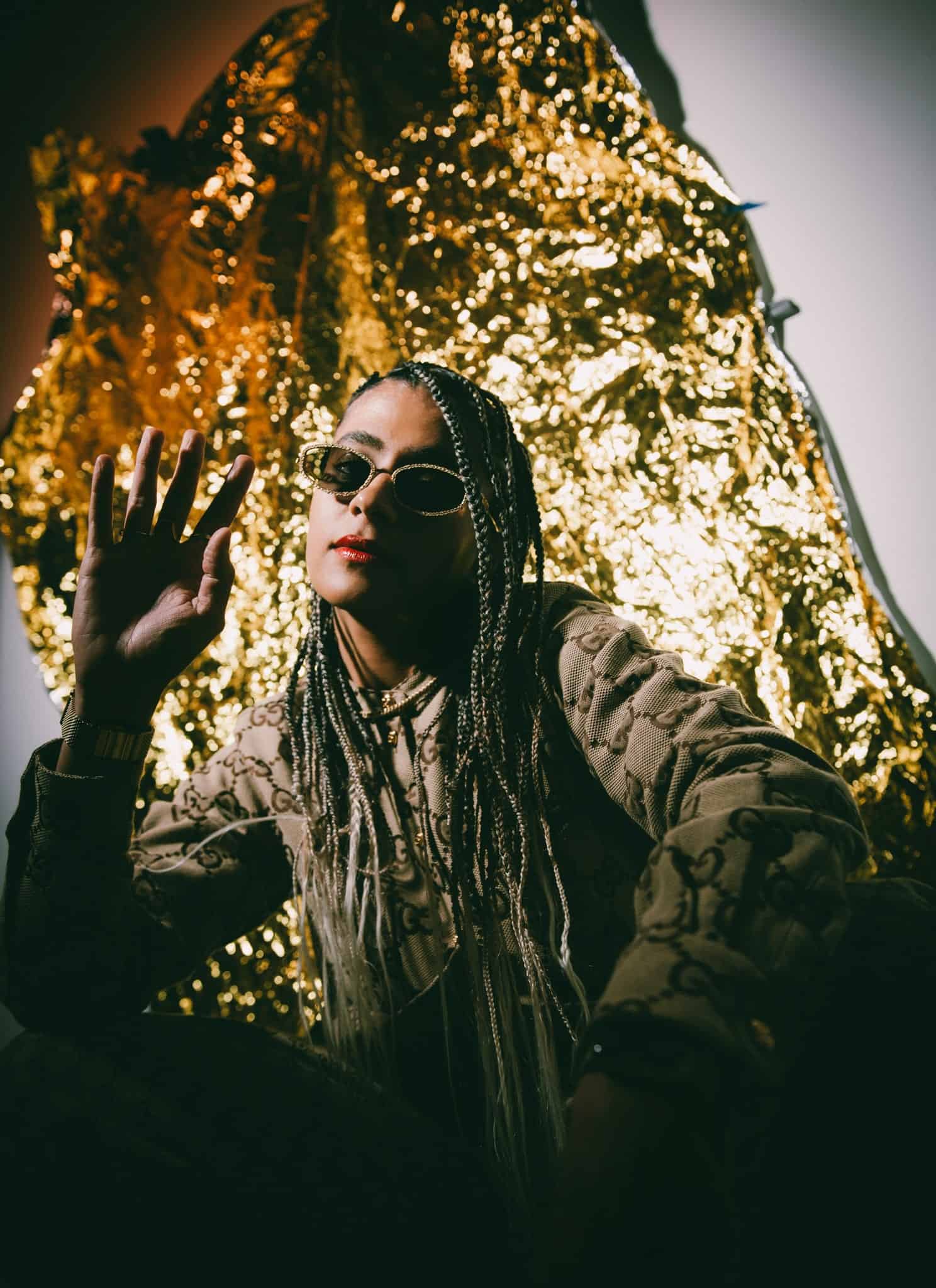 Singer Madame Gandhi, with long black and blonde braids, and sunglasses, against a gold backdrop.