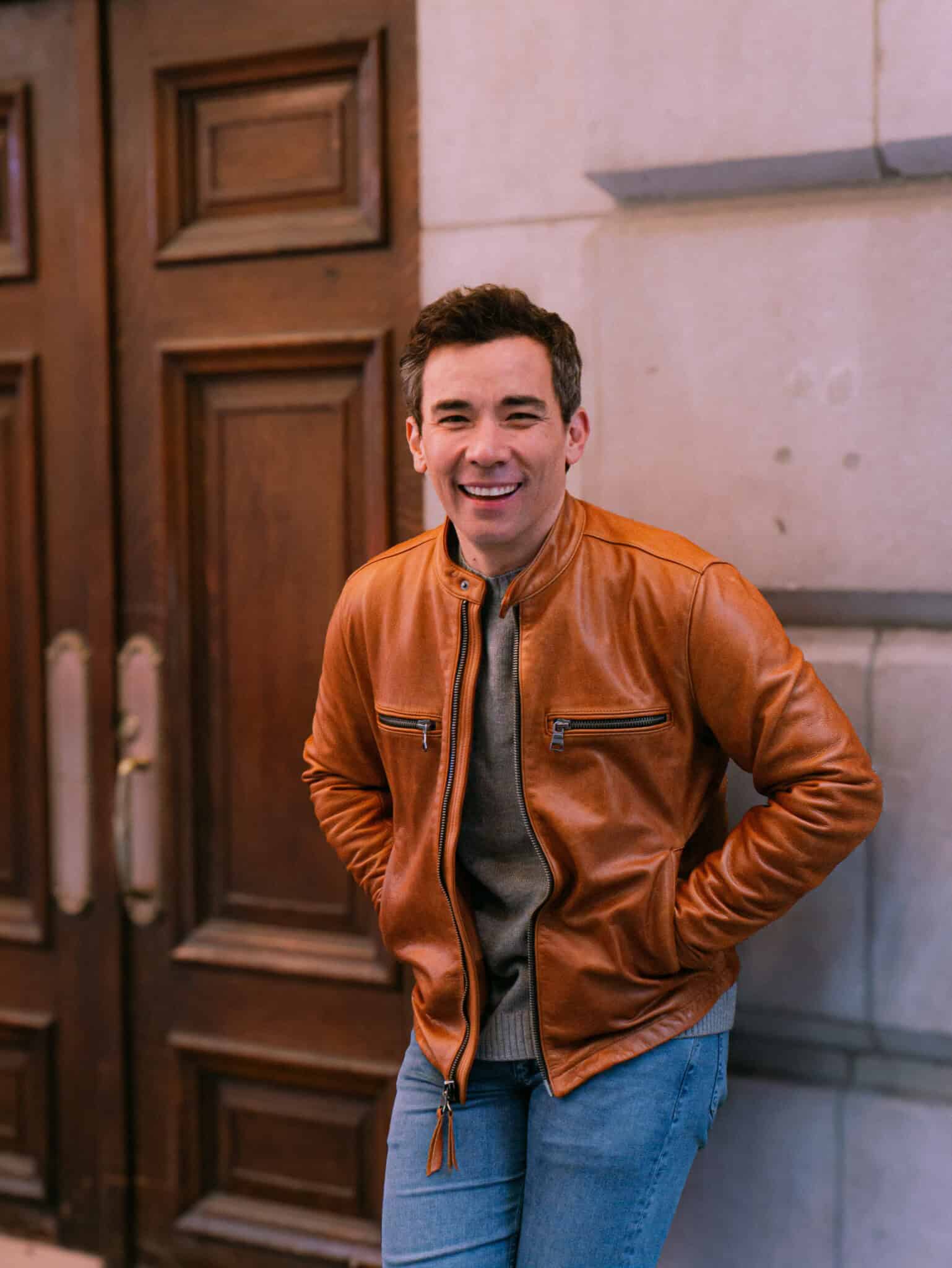 Conrad Ricamora stands against a wall smiling.