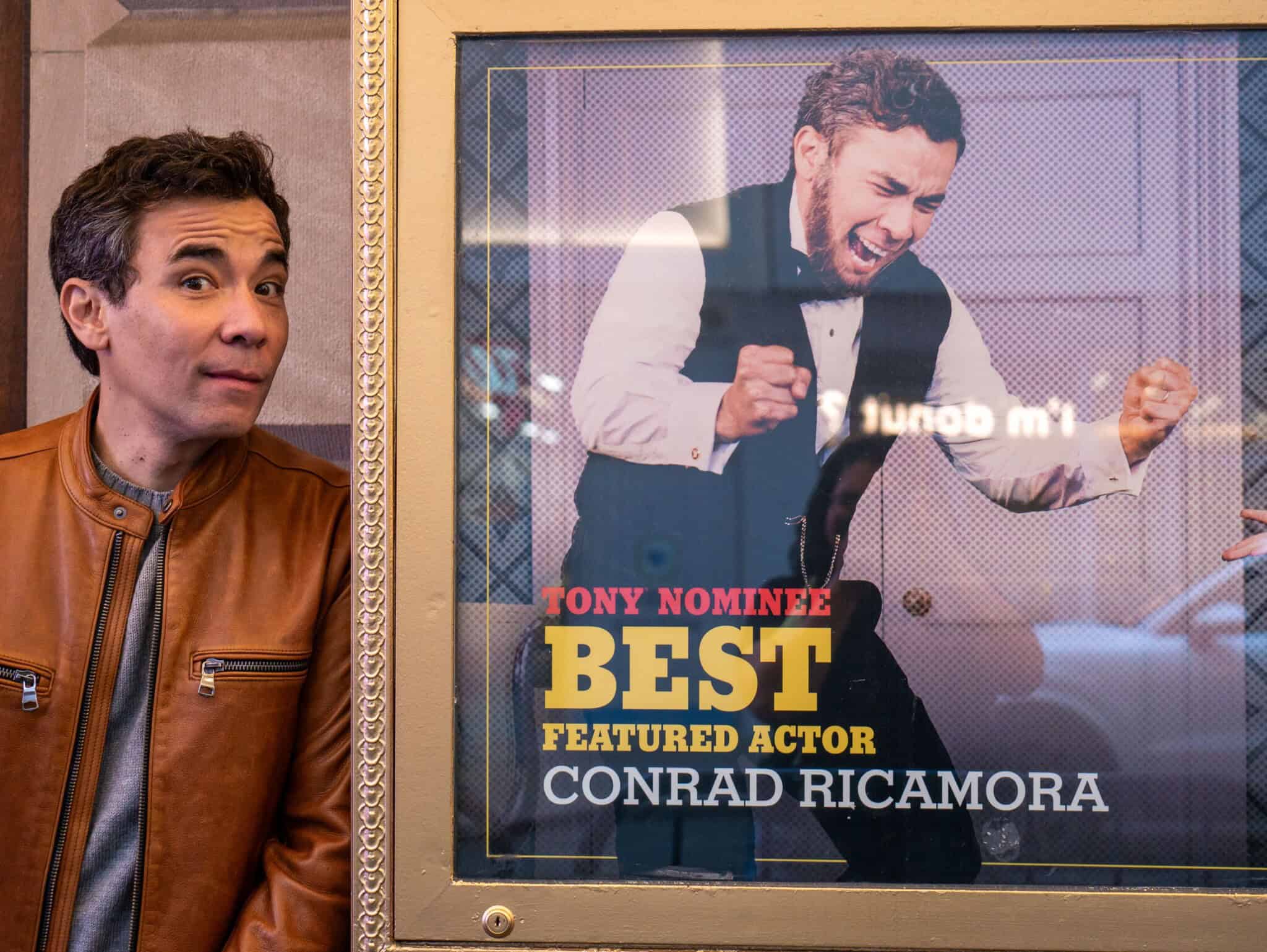 Conrad Ricamora stands to the left of a photo of himself in character as Lincoln that reads “Tony Nominee Best Featured Actor - Conrad Ricamora”