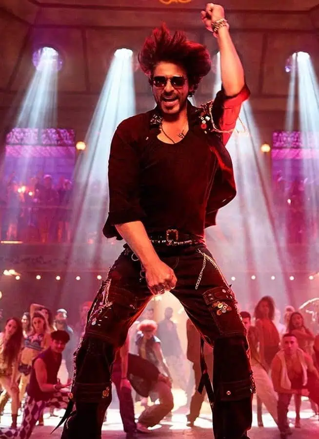 Actor Shah Rukh Khan, dressed in black, with black sunglasses, dances, with a crowd behind him.