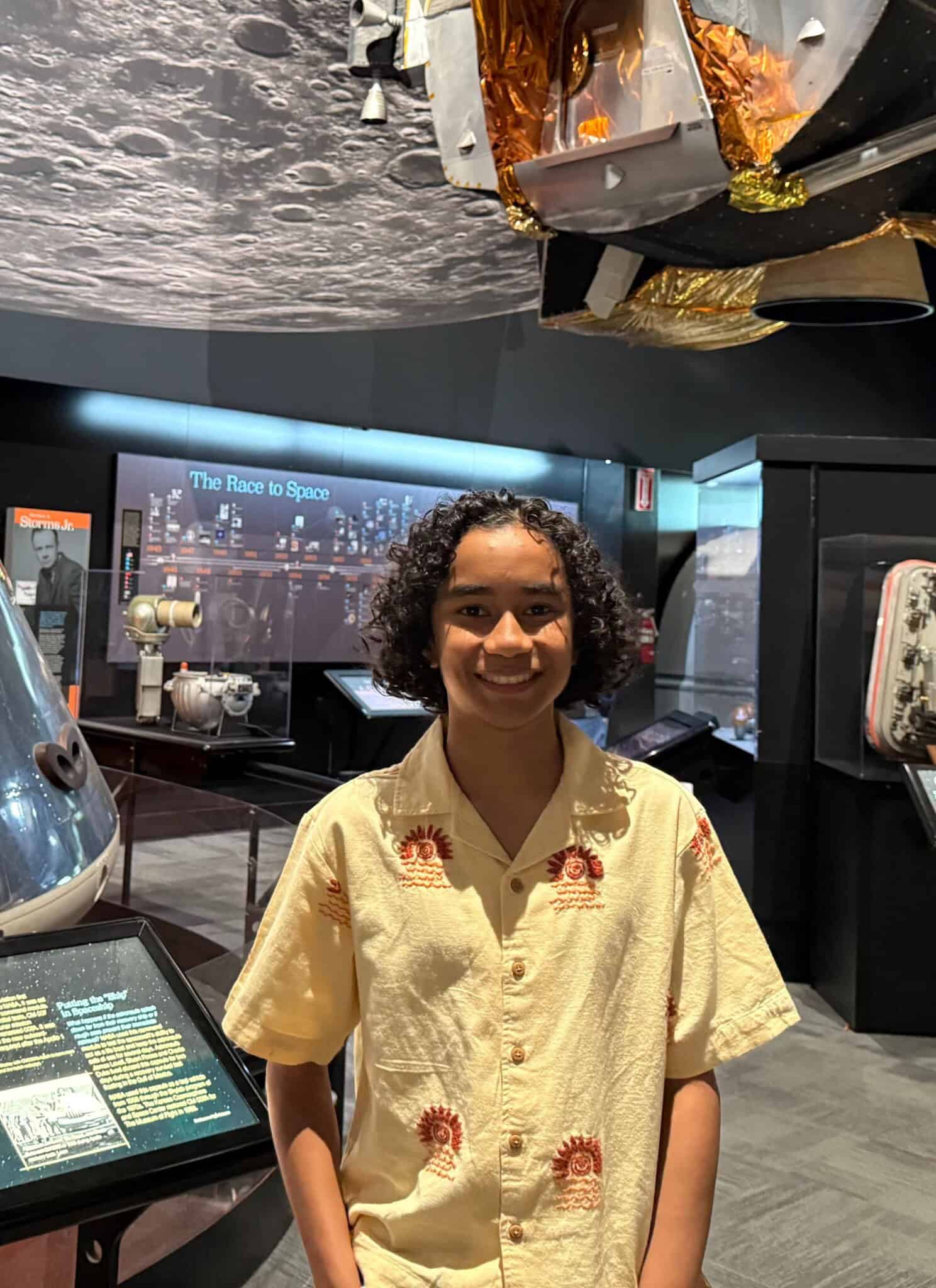 A teenage boy with curly black hair, in a light cream top, stands in front of museum displays about space.