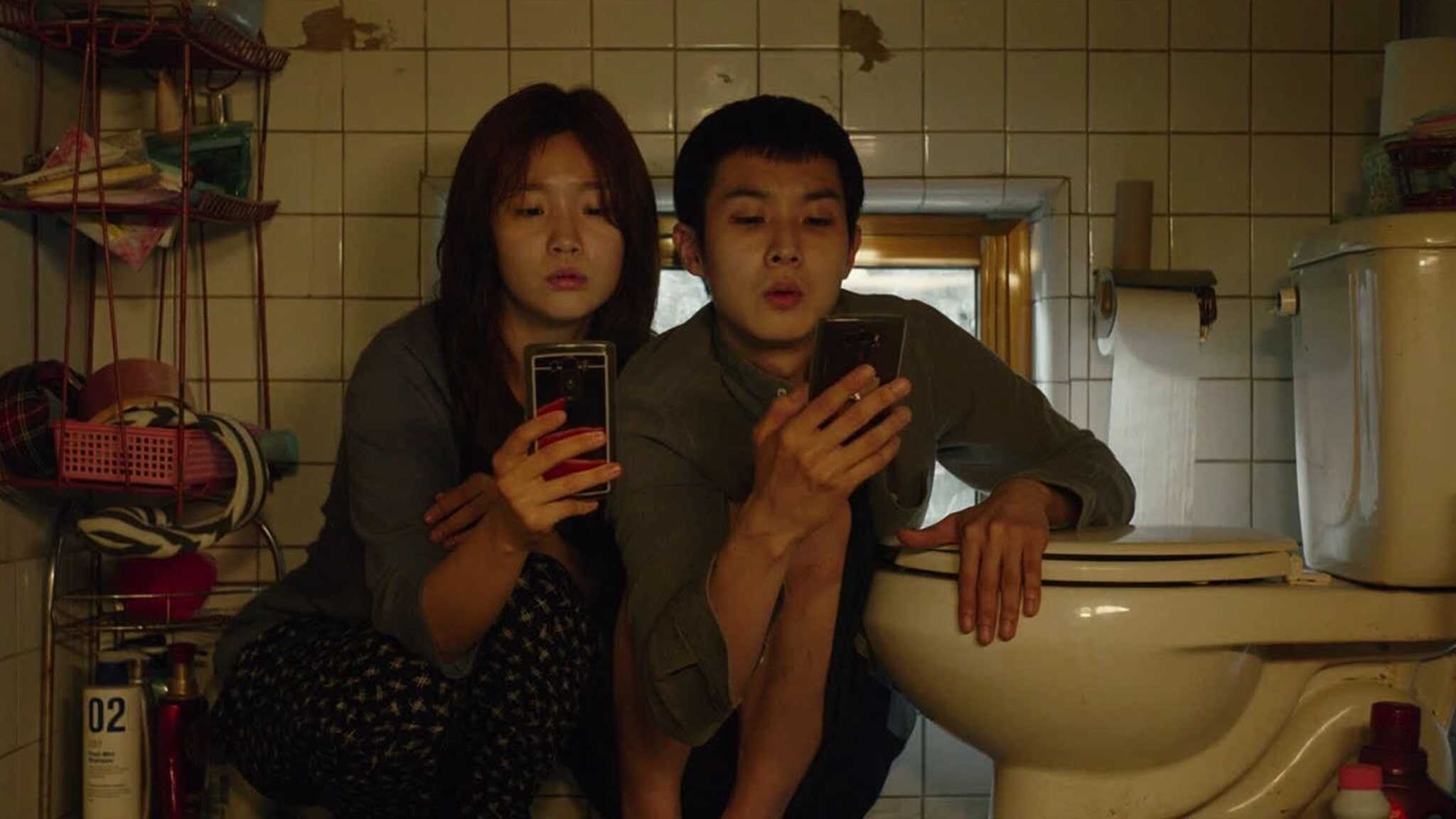 Actors Park So-dam and Choi Woo-shik sit together, looking at cell phones next to a toilet in 