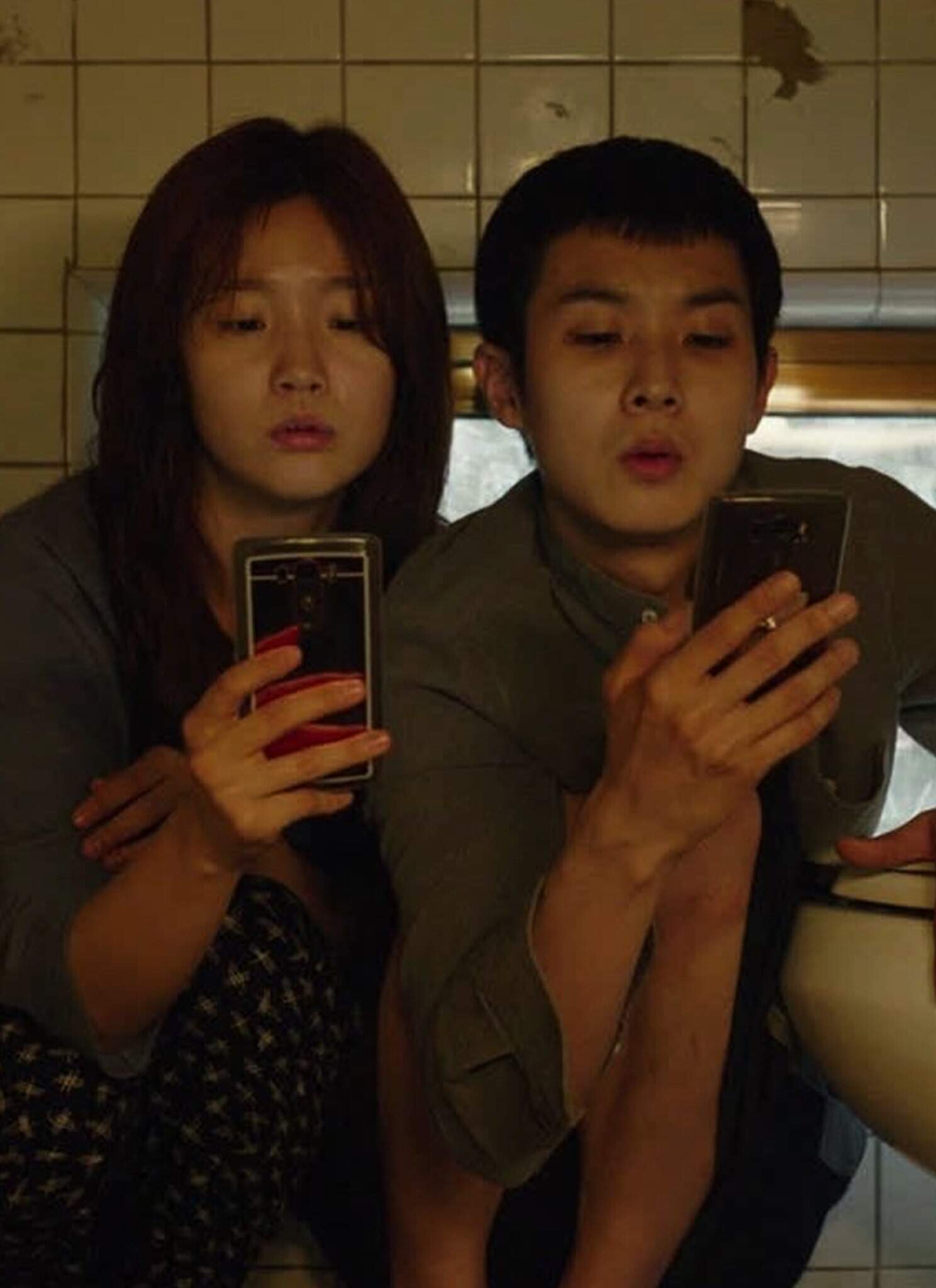 Actors Park So-dam and Choi Woo-shik sit together, looking at cell phones next to a toilet in "Parasite."