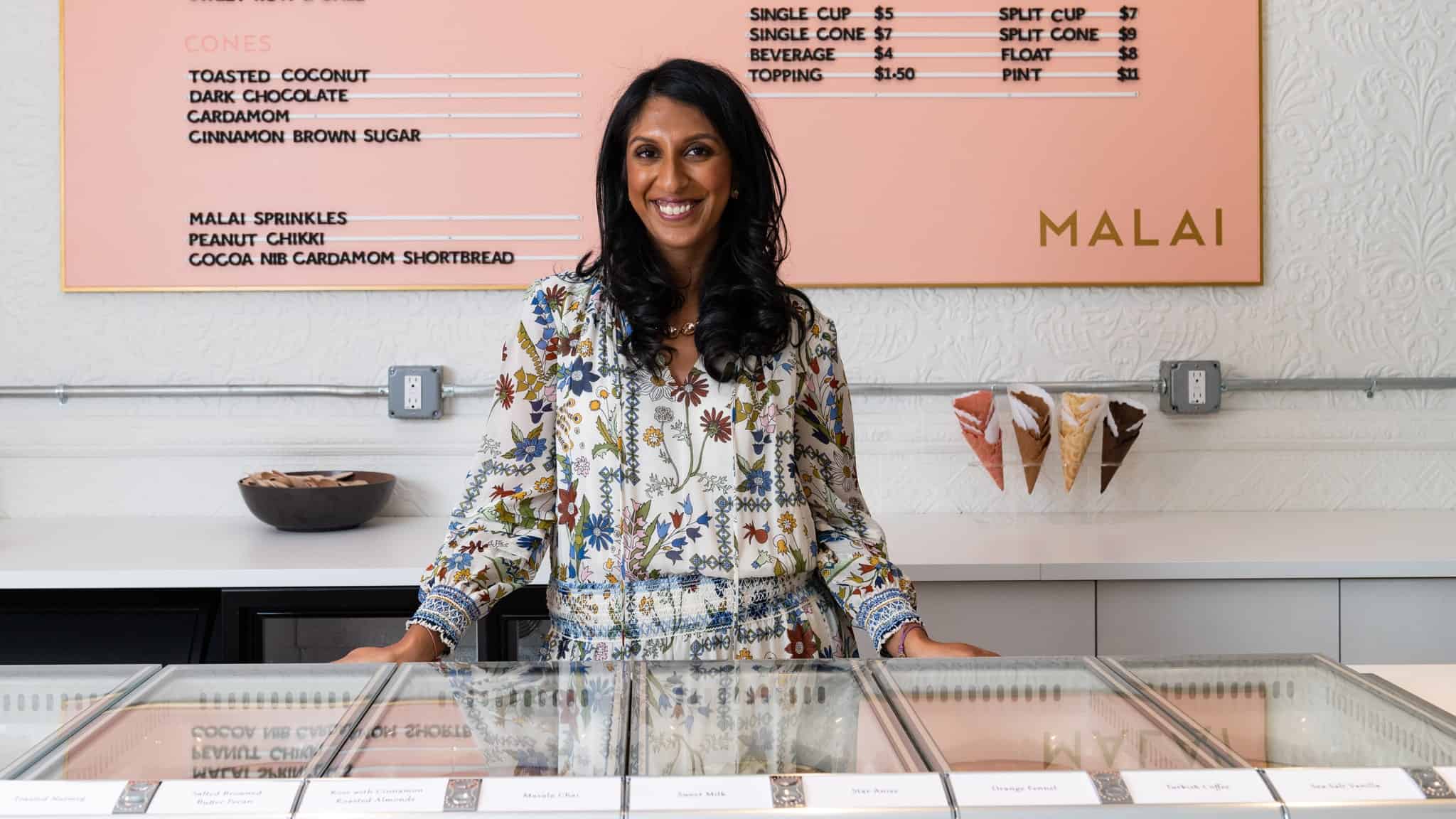 A South Asian woman in a white floral dress stands at an ice cream counter with a pink price list behind her.