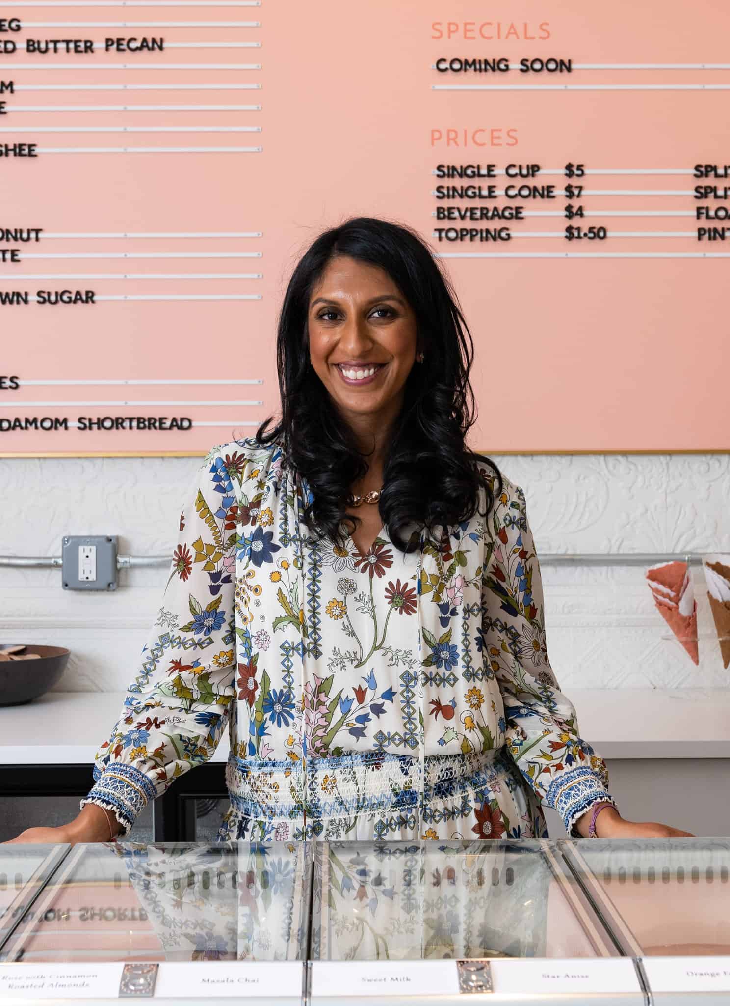A South Asian woman in a white floral dress stands at an ice cream counter with a pink price list behind her.