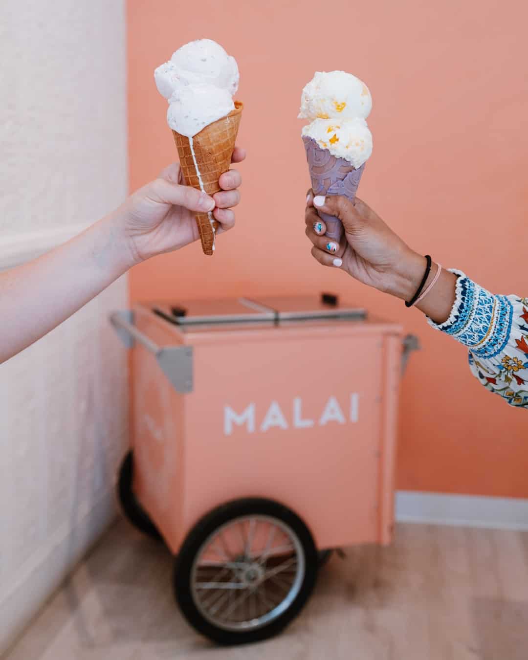 Two hands hold up ice cream cones, with a pink cart with "Malai" on the front, in the background.