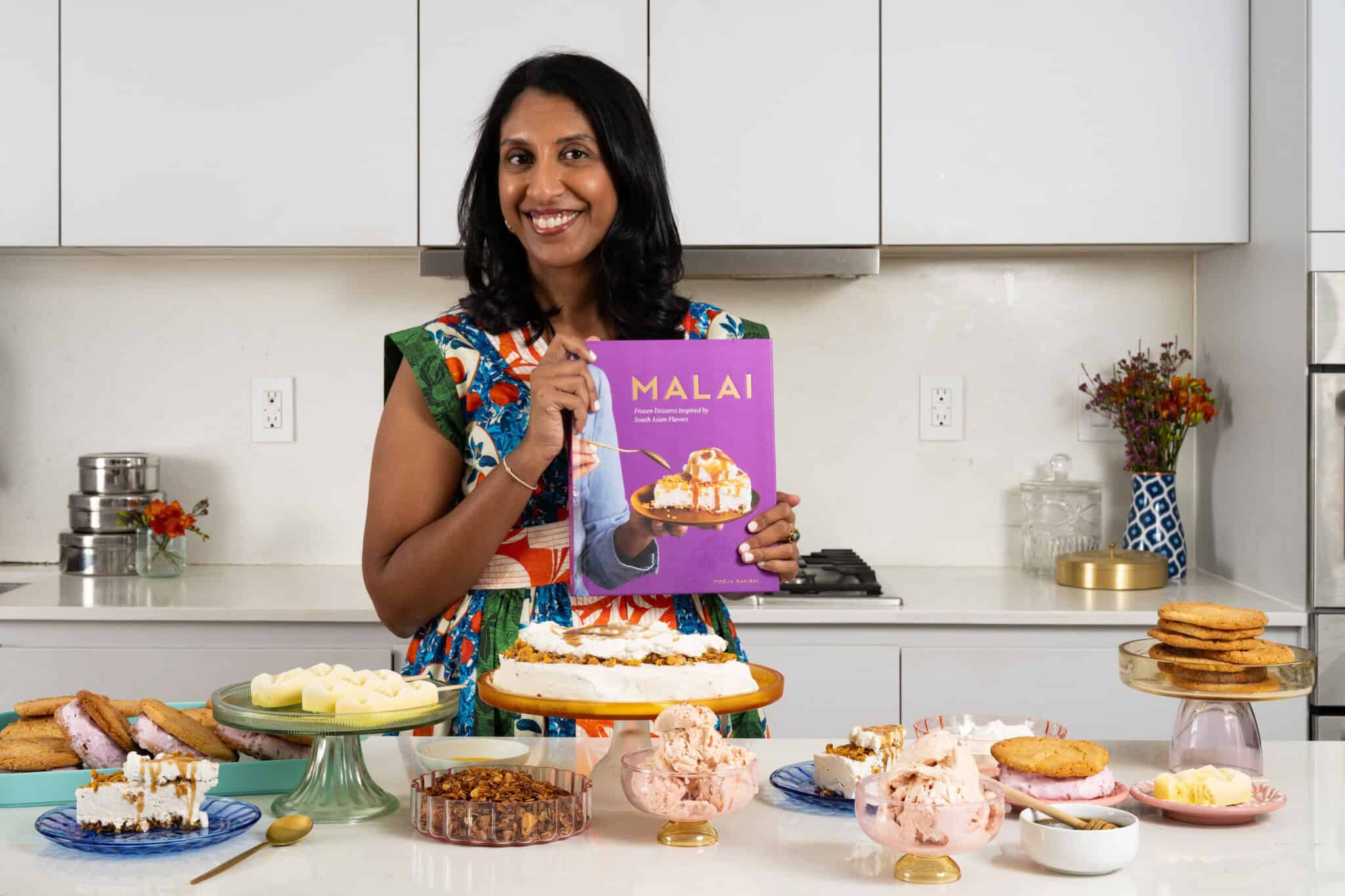 A South Asian woman stands in a kitchen with desserts on the counter, holding a book titled, "Malai."