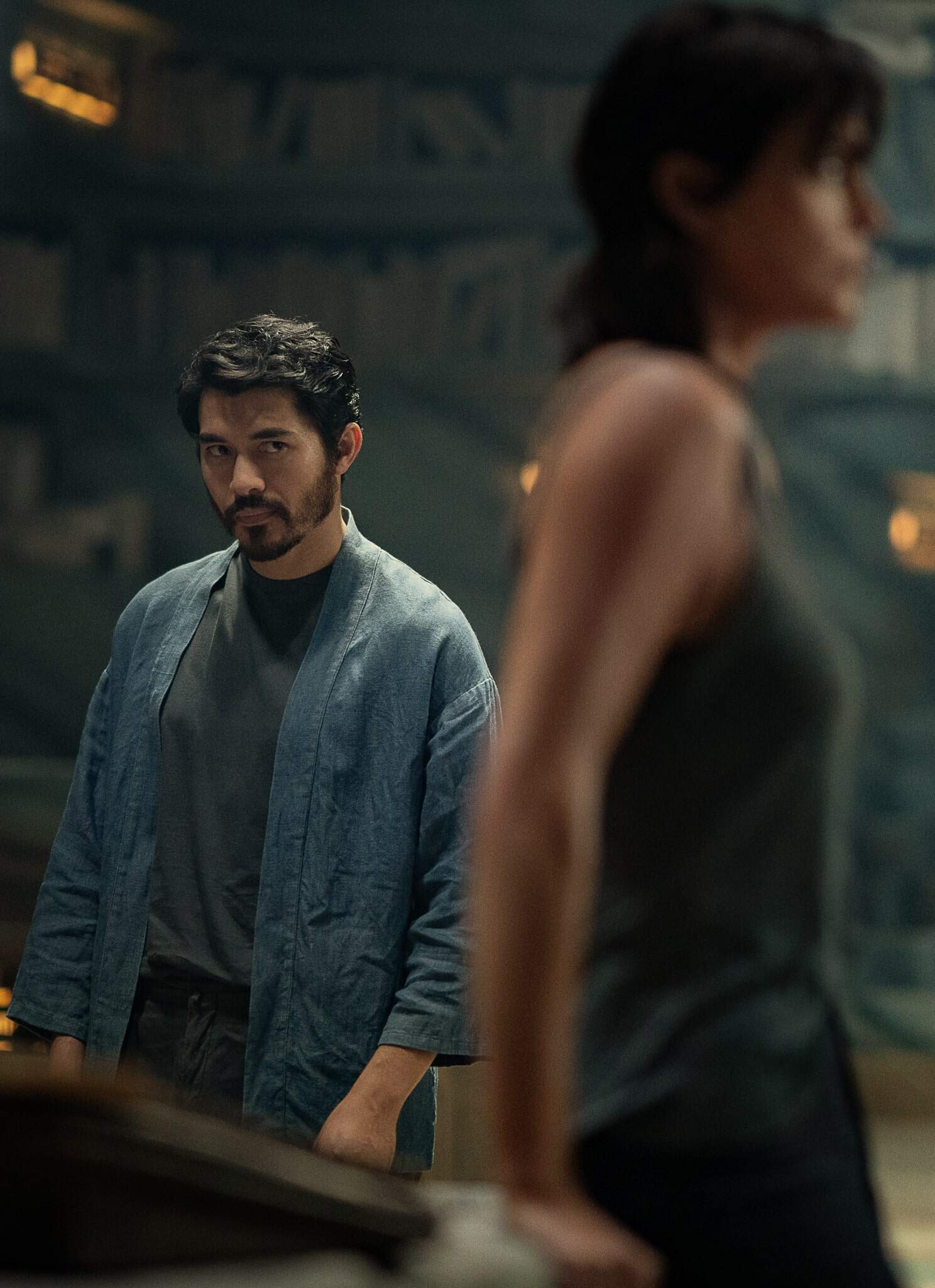Actor Henry Golding stands in the background in a blue kimono top, with actress a blurred out Charlize Theron in the foreground in "The Old Guard 2."