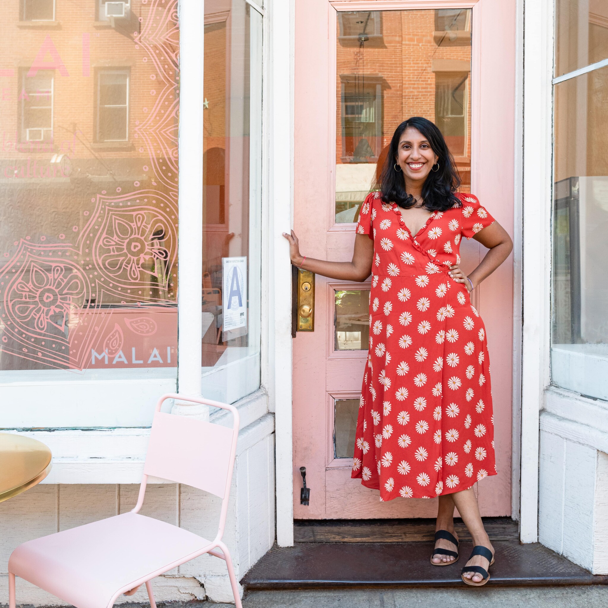 A South Asian woman in a read floral dress stands in a doorway with a pink door behind her.