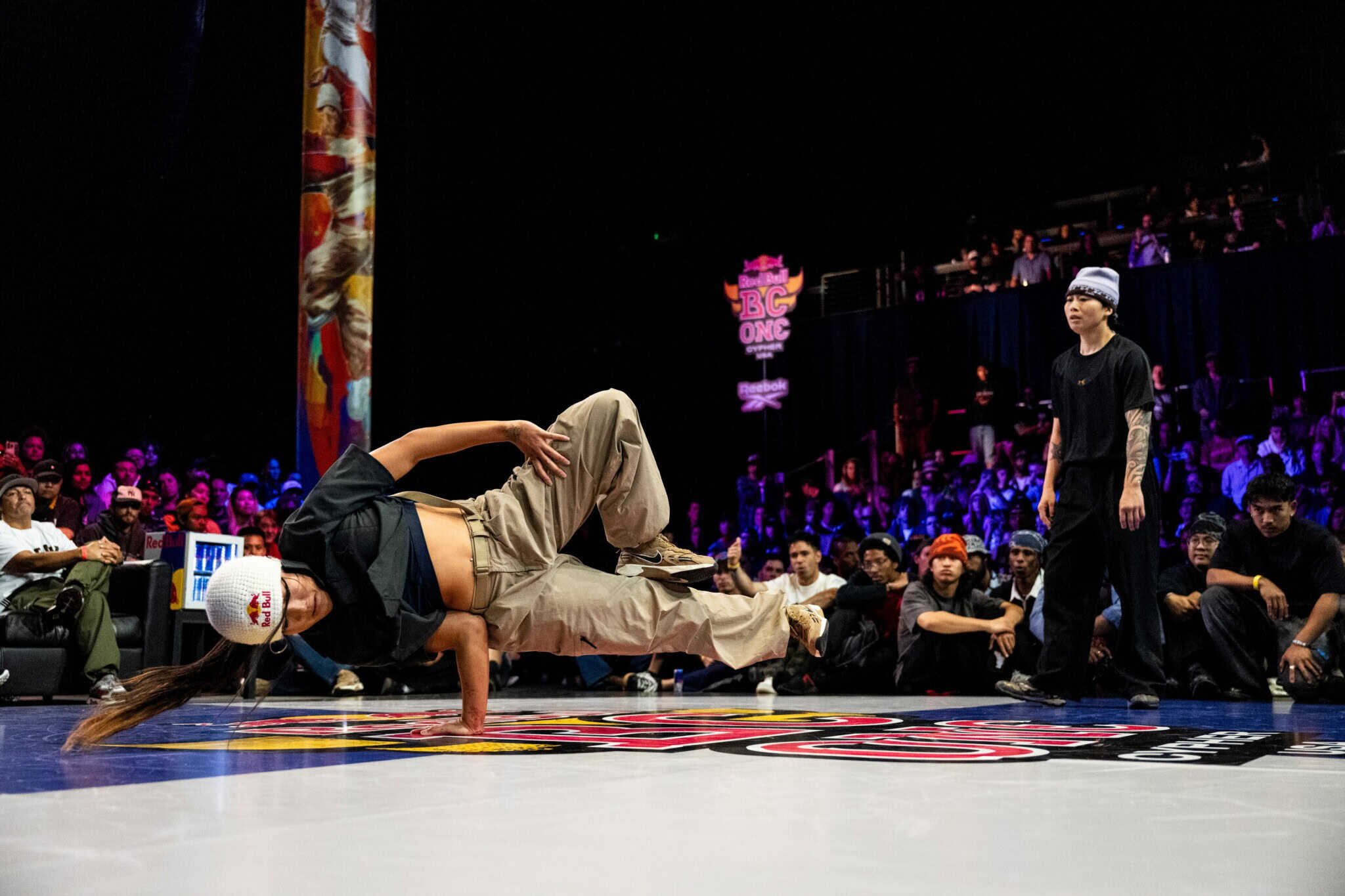 A breakdancer in a black top, khaki pants and white beanie balances on one arm, with a crowd in the background.