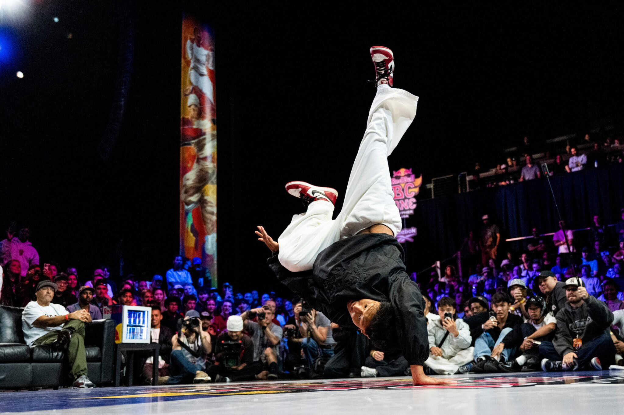 A breakdancer in a black top and white pants poses upside down on one hand, with a crowd in the background.