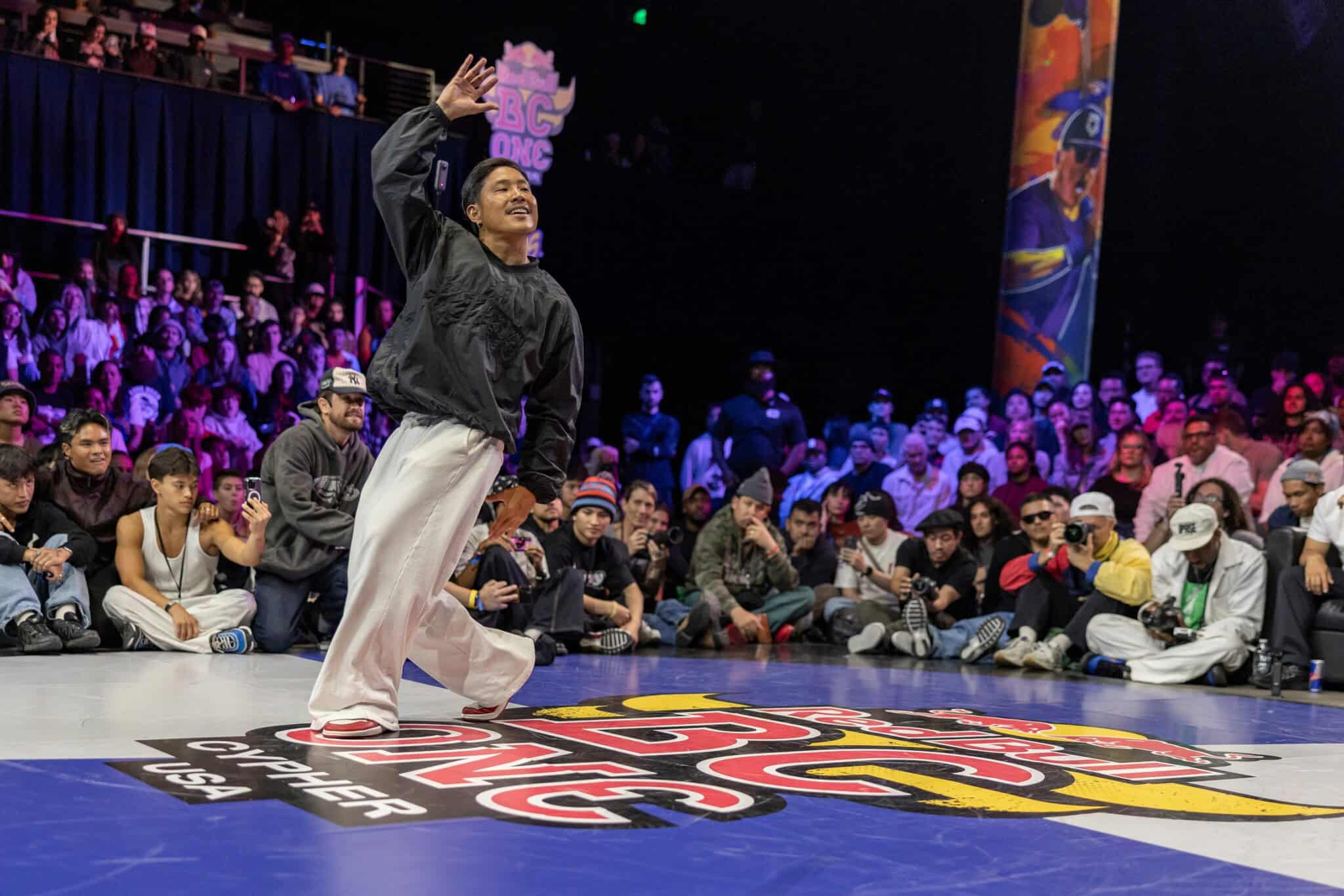 A breakdancer in a black top and white pants stands with his arm up, with a crowd in the background.