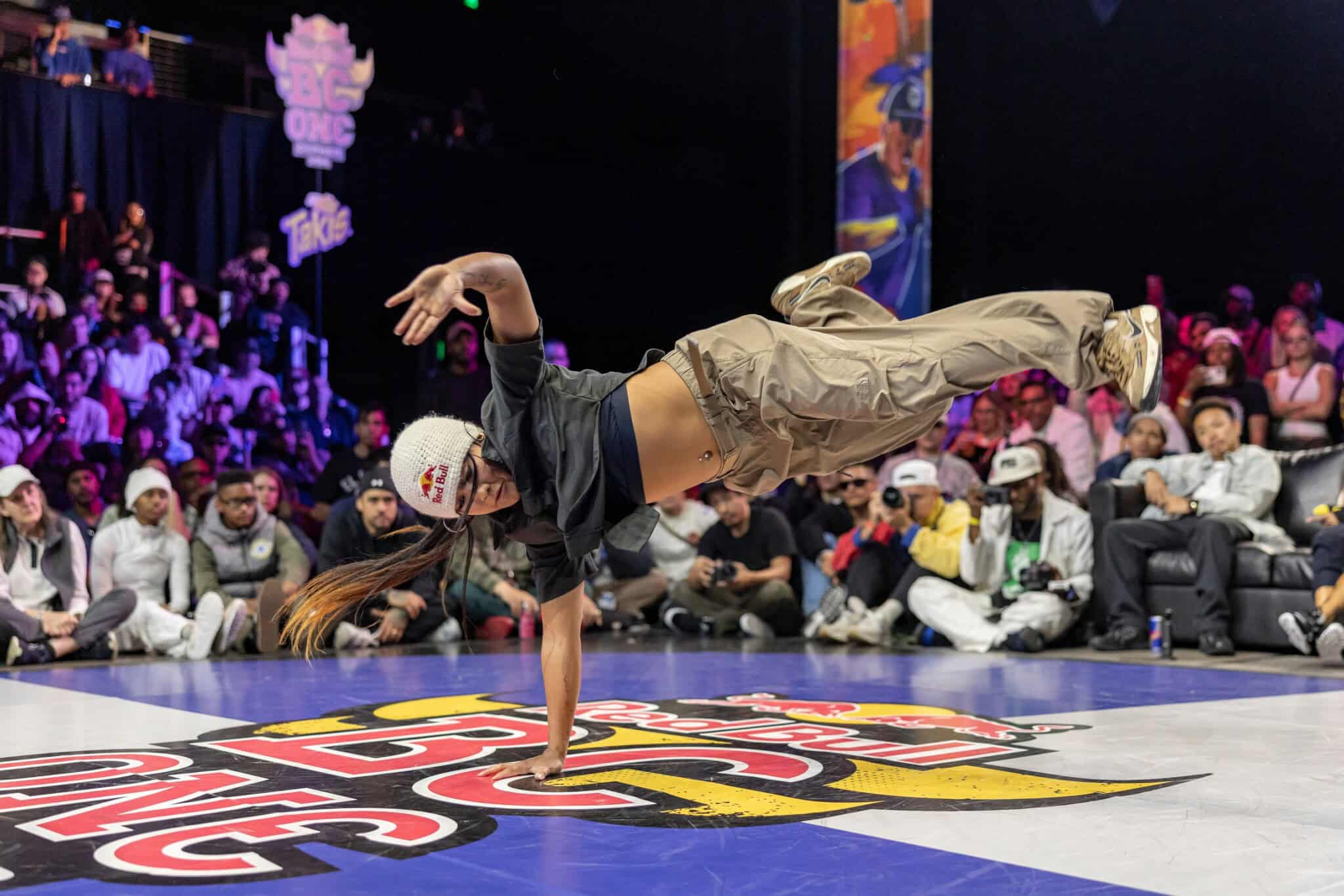 A breakdancer in a black top, khaki pants and white beanie balances on one arm, with a crowd in the background.