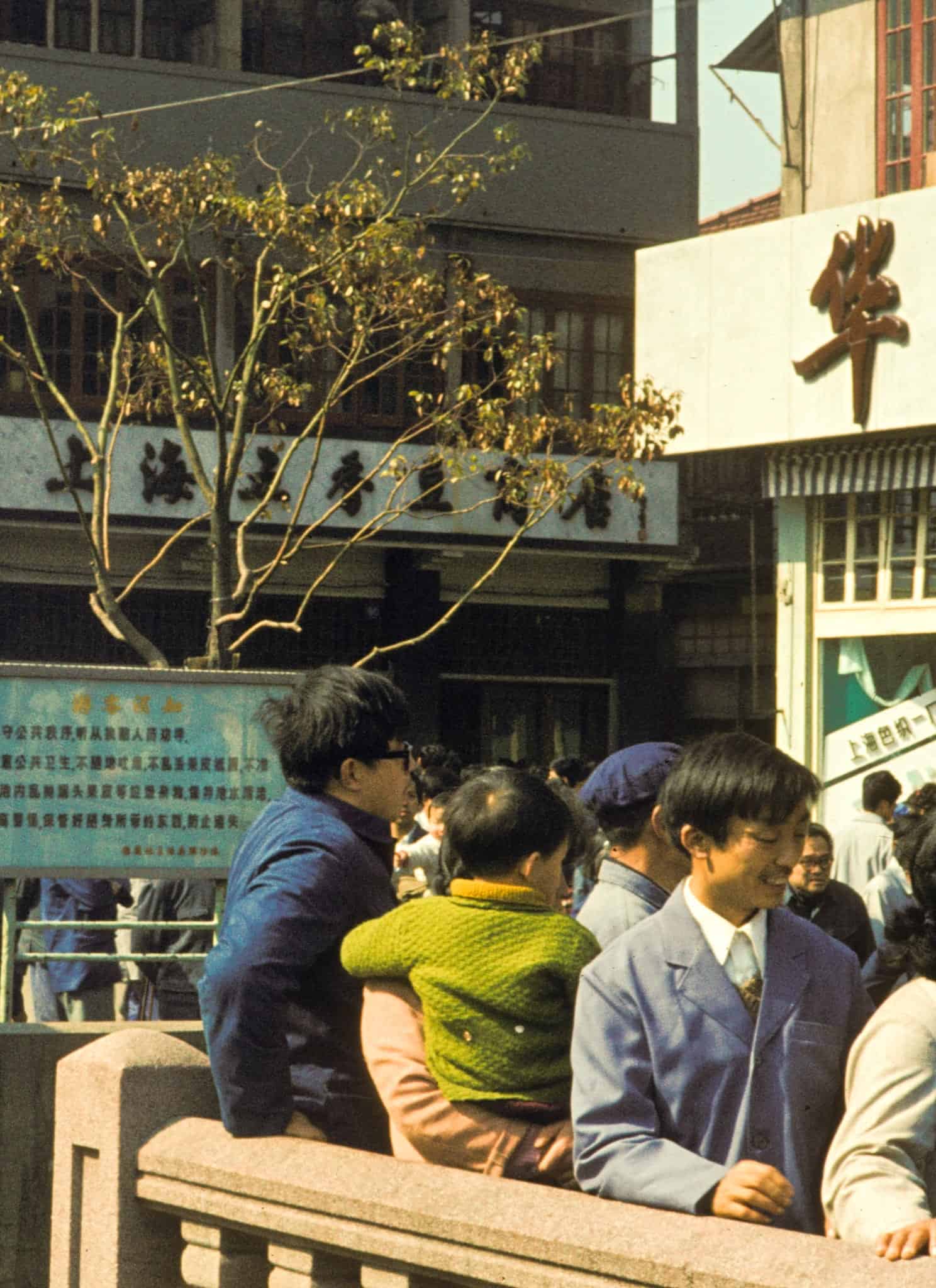 A group of Chinese people stand outside business buildings against a stone railing in China in 1982.