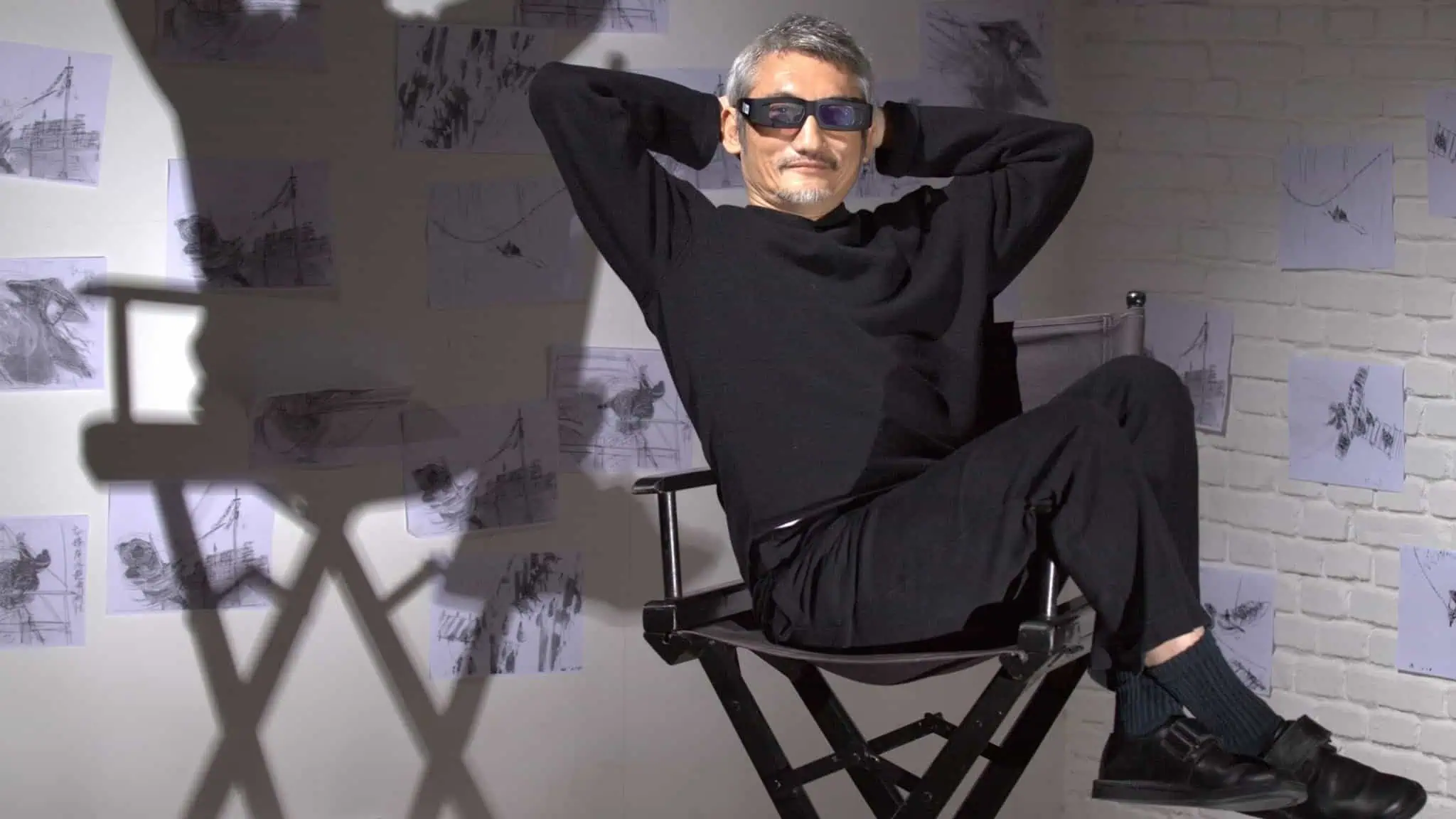 Filmmaker Tusi Hark sits in a director's chair with his hands behind his head, dressed in black and sunglasses, with his shadow behind him.