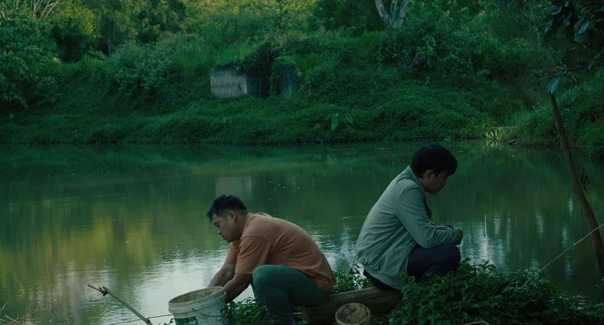 Two Asian men sit with their backs to each other in front of a body of water in "Inside the Yellow Cocoon Shell."