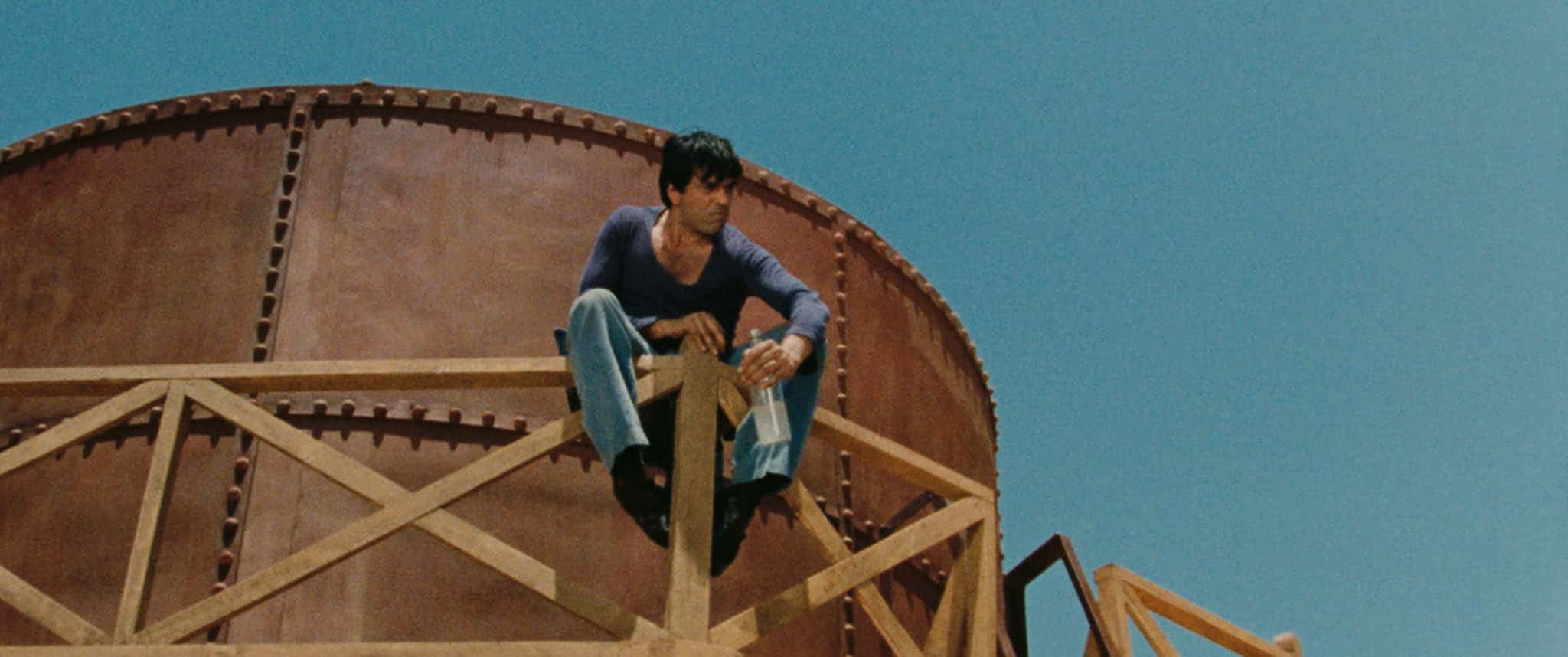 A man in a blue shirt and jeans crouches on a wooden railing in front of a large, round metal structure under a clear blue sky.