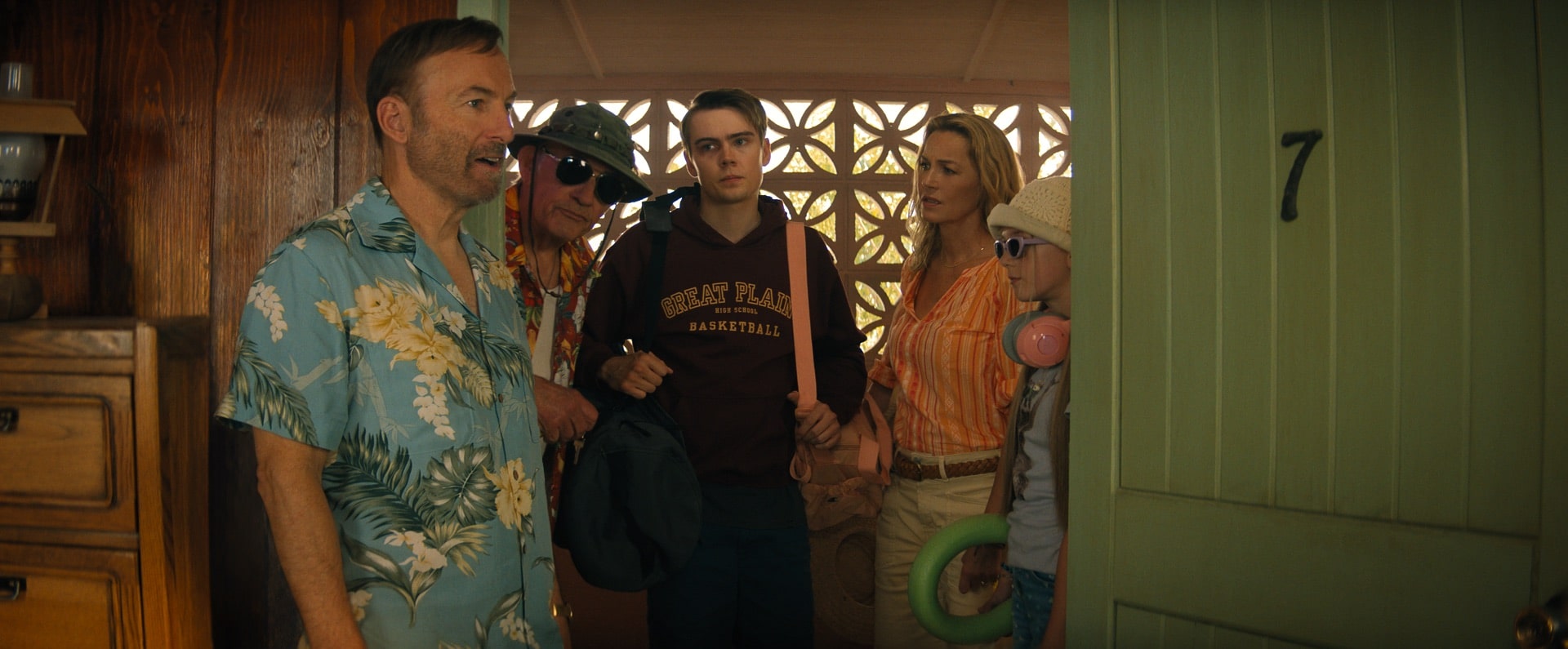 A man in a blue floral shirt stands by an open green door marked 7, as four people with luggage, sunglasses, and swim gear appear ready for vacation. The group looks inside with mixed expressions.