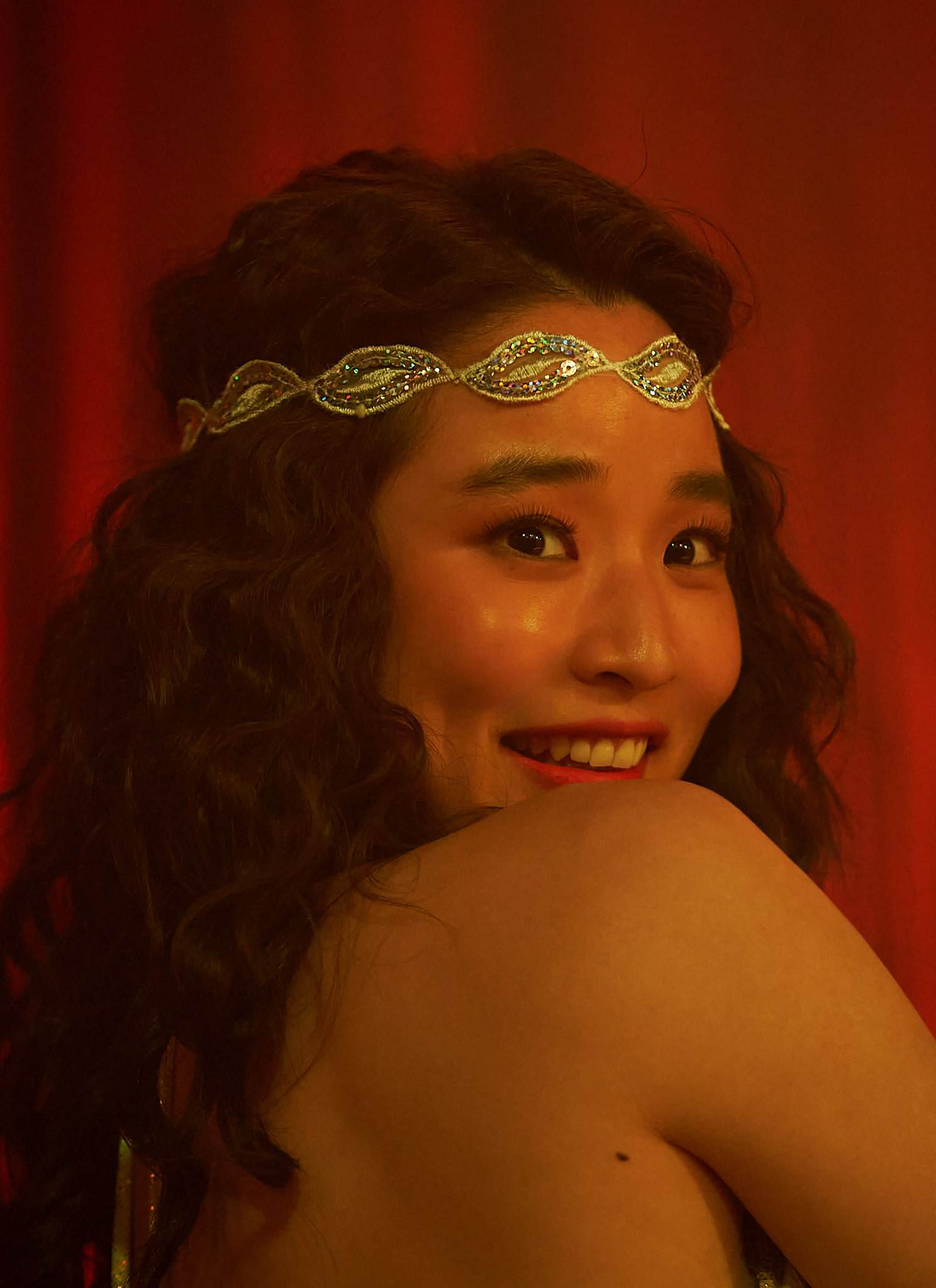 A woman with curly hair, wearing a jeweled headband and shimmering green costume, smiles over her shoulder in front of red curtains, clapping her hands together.