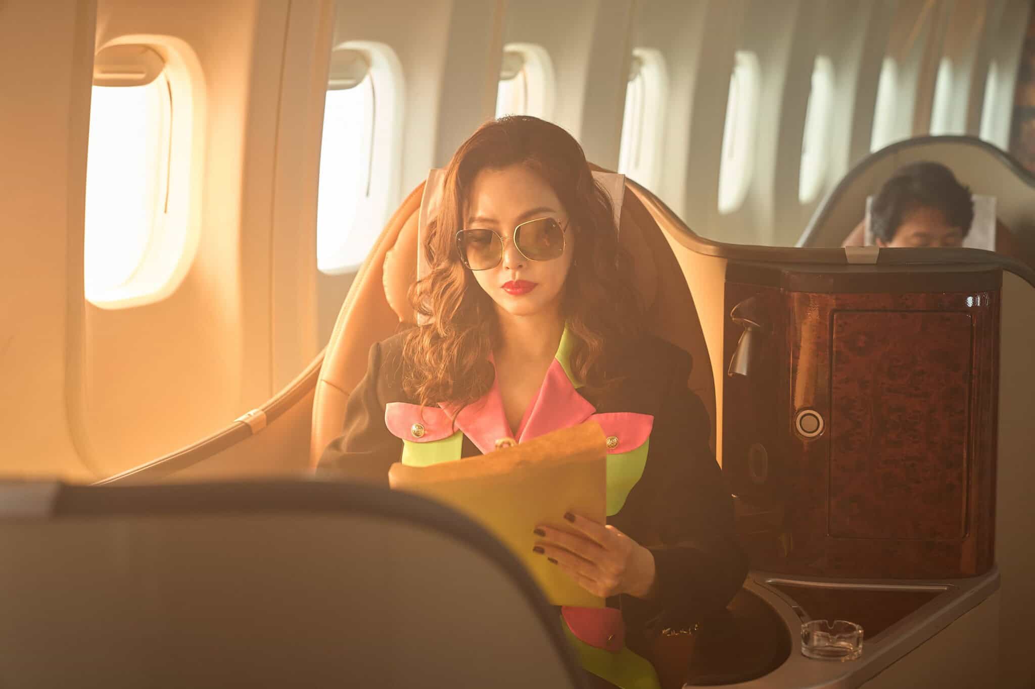 A woman with wavy hair and sunglasses sits in a first-class airplane seat, reading a document. She wears a stylish black and pink outfit, and sunlight streams through the nearby windows.