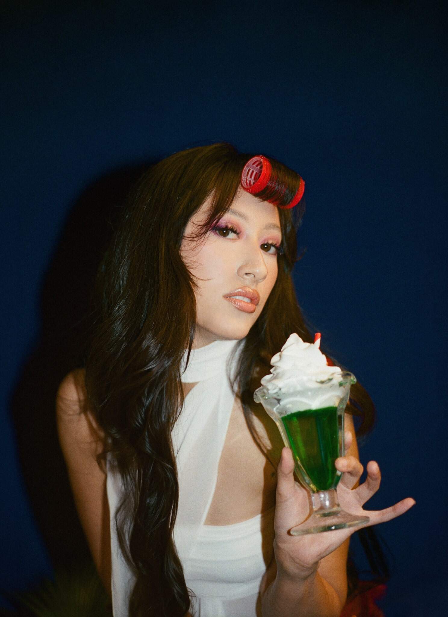 A woman with long dark hair and a red hair roller in her bangs holds a glass of green dessert topped with whipped cream and a cherry, posing against a dark blue background.