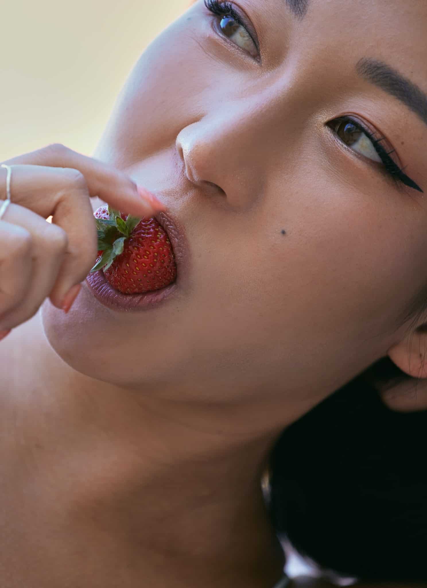 A close-up of a woman with winged eyeliner and a lace top, lying down and holding a strawberry near her lips as she prepares to take a bite.