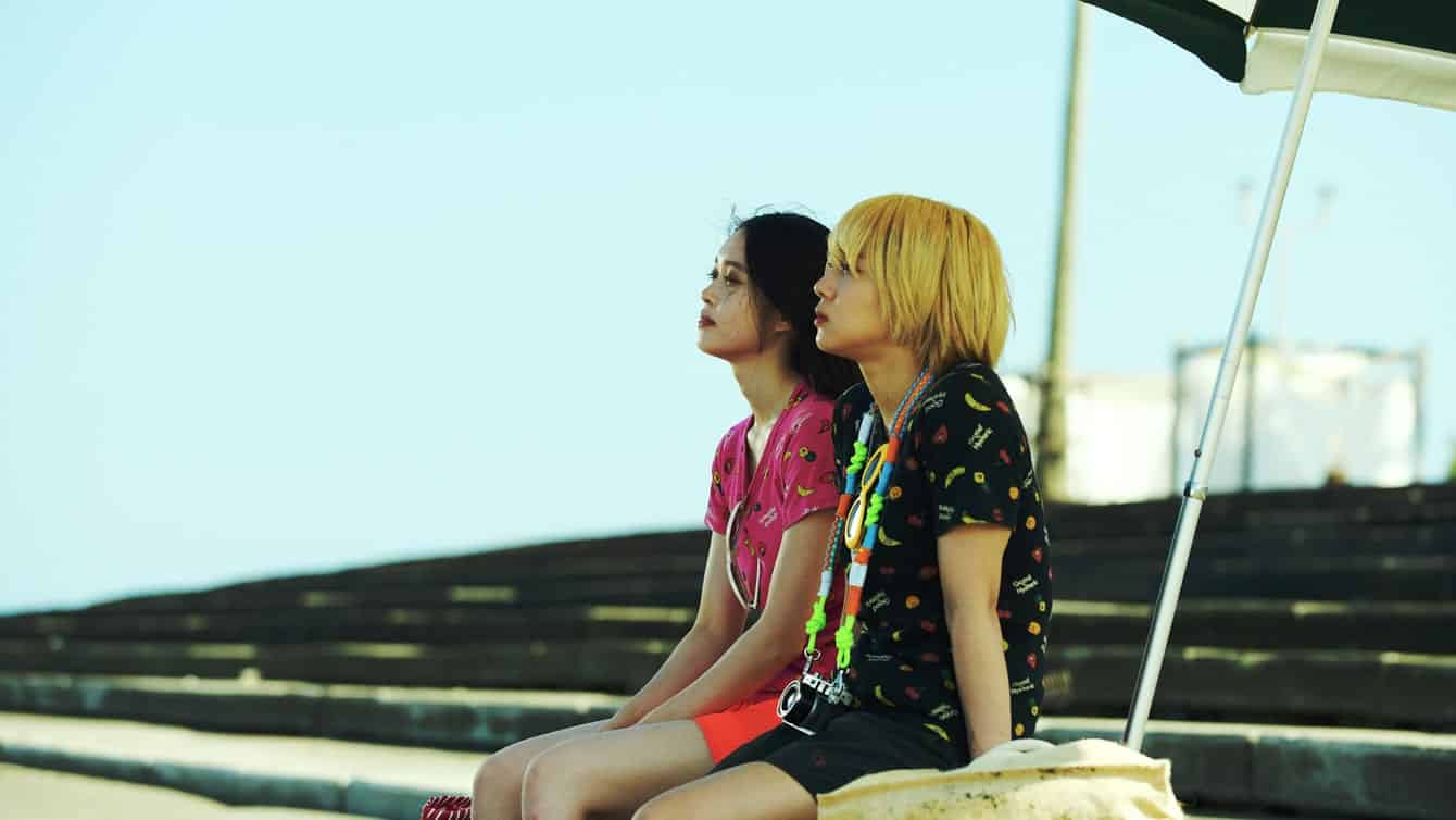 Two young people sit side by side on a stone ledge under an umbrella, looking thoughtful. One has short blond hair and colorful clothes, the other has long dark hair and a pink shirt. Steps and a blue sky are in the background.