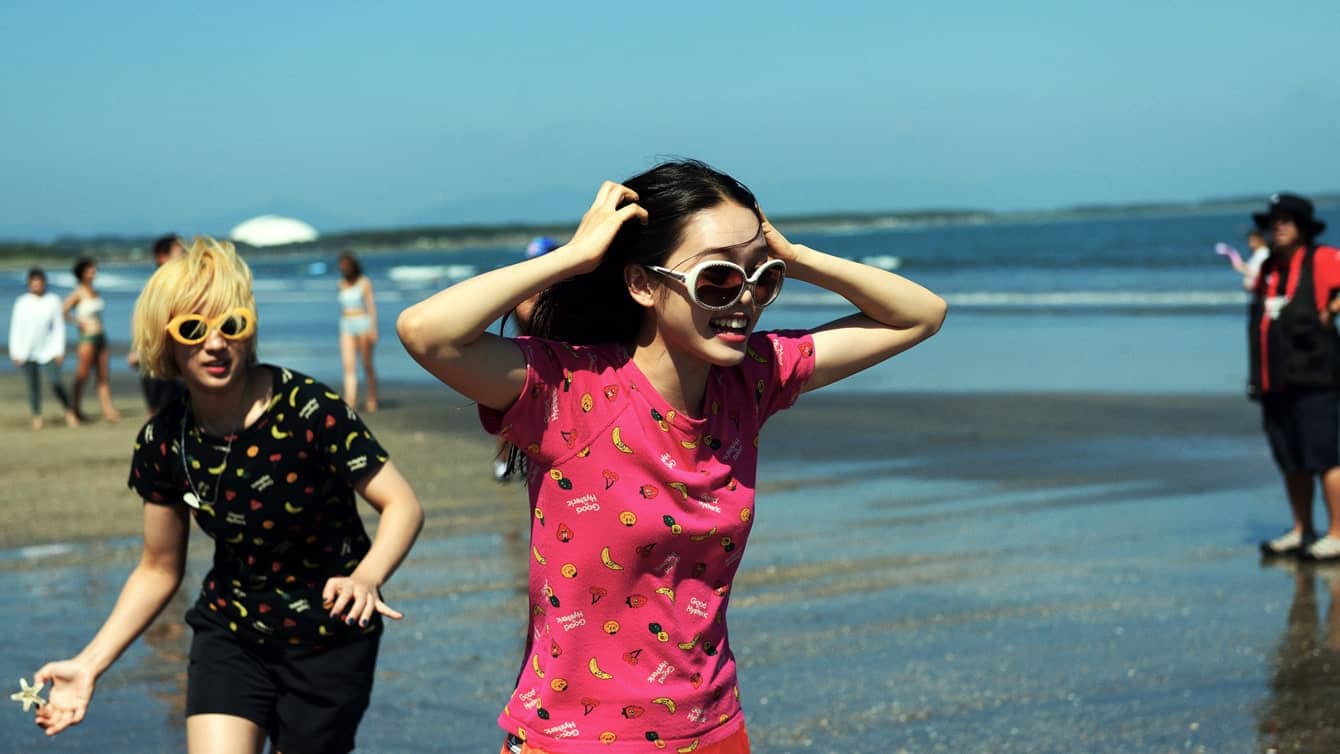 Two young women wearing sunglasses and colorful shirts smile and run along a sunny beach. People and the ocean are in the background under a clear blue sky.