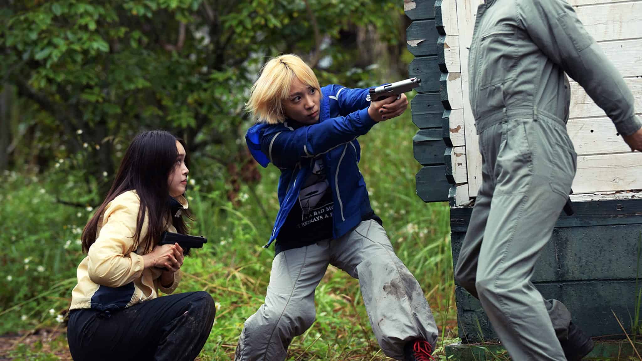Two people with guns take cover beside a wooden building in a grassy outdoor area, aiming at a person in gray running around the corner. The scene looks tense and action-packed.