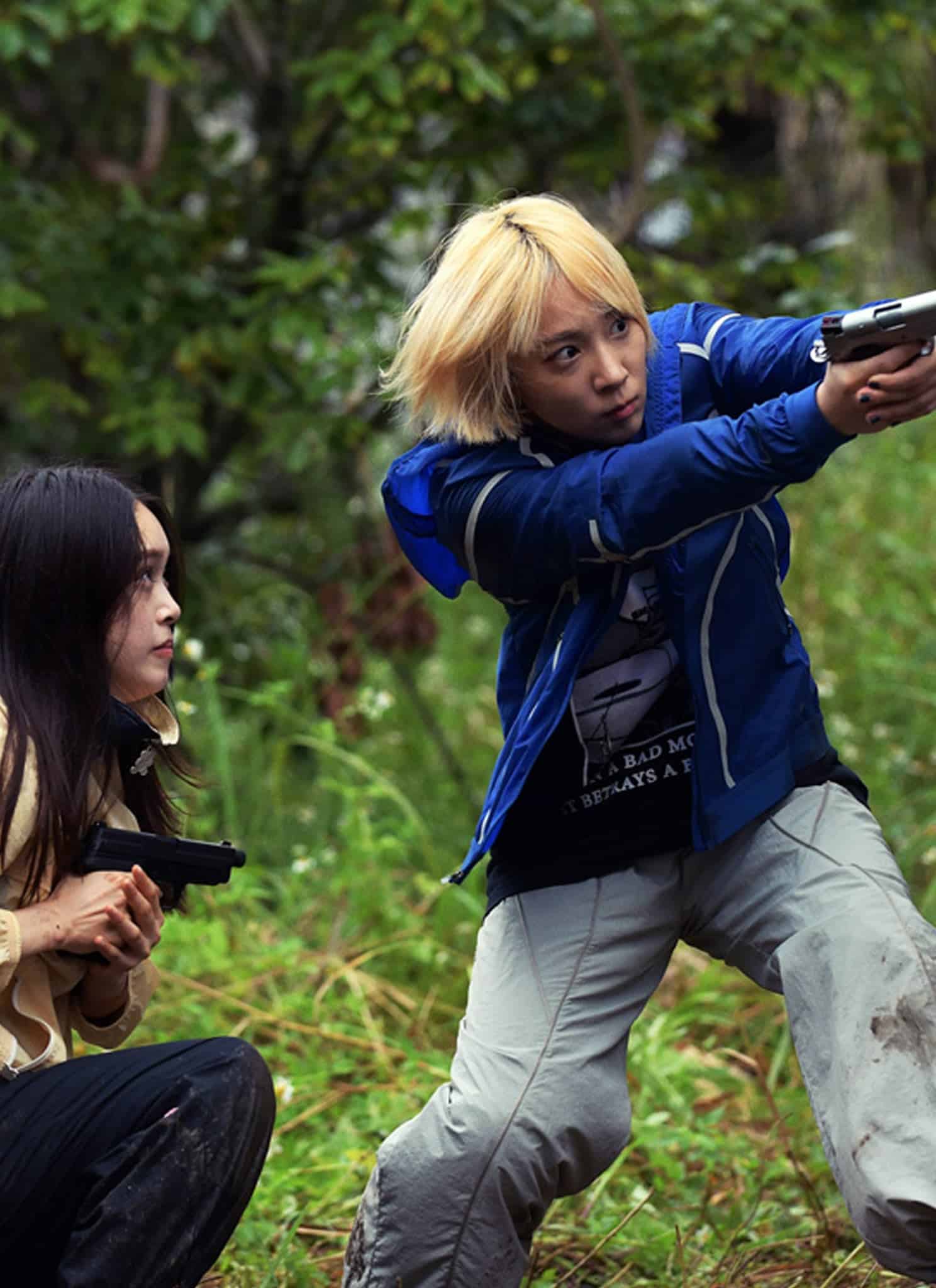 Two people with guns take cover beside a wooden building in a grassy outdoor area, aiming at a person in gray running around the corner. The scene looks tense and action-packed.