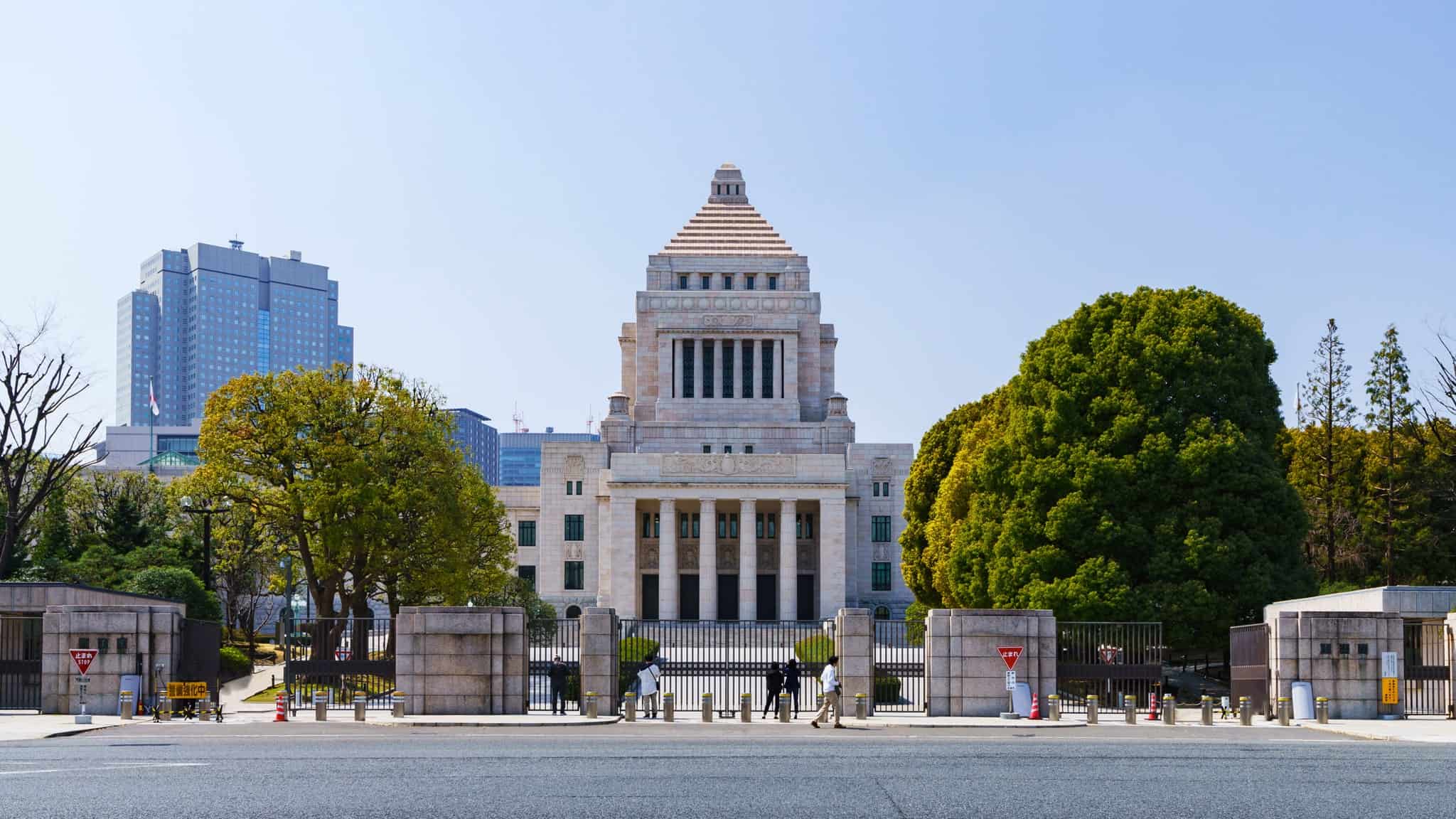 The image shows the front view of Japan’s National Diet Building in Tokyo, symbolizing Japanese conservatism with its distinctive pyramid-shaped roof, flanked by trees and modern buildings. People are walking near the entrance gate.