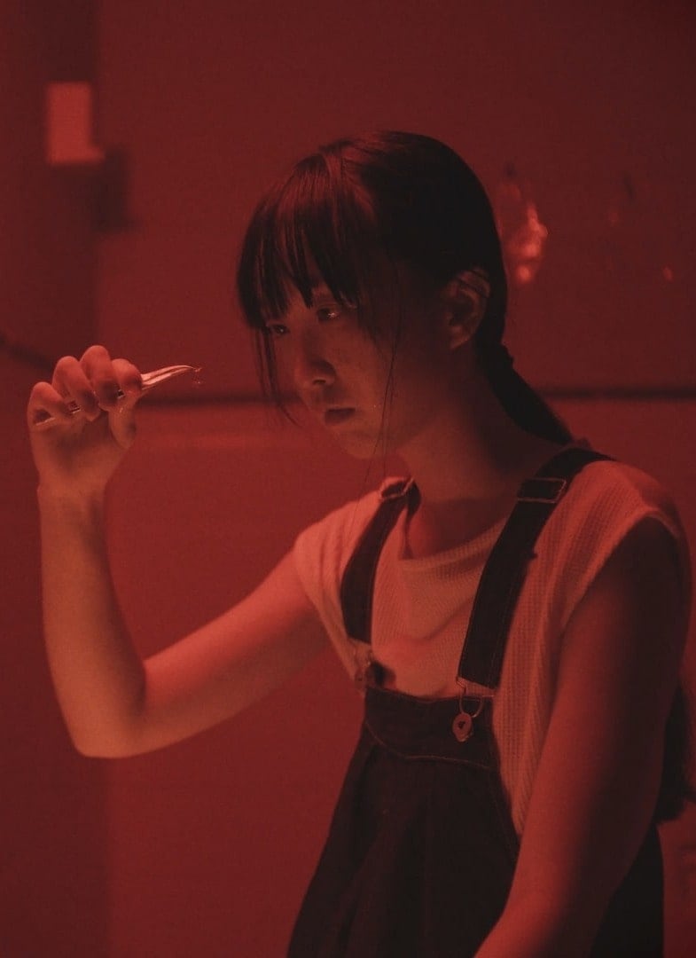 A young person in overalls holds up tweezers holding a contact lens, under red lighting, looking at it intently with a serious expression.