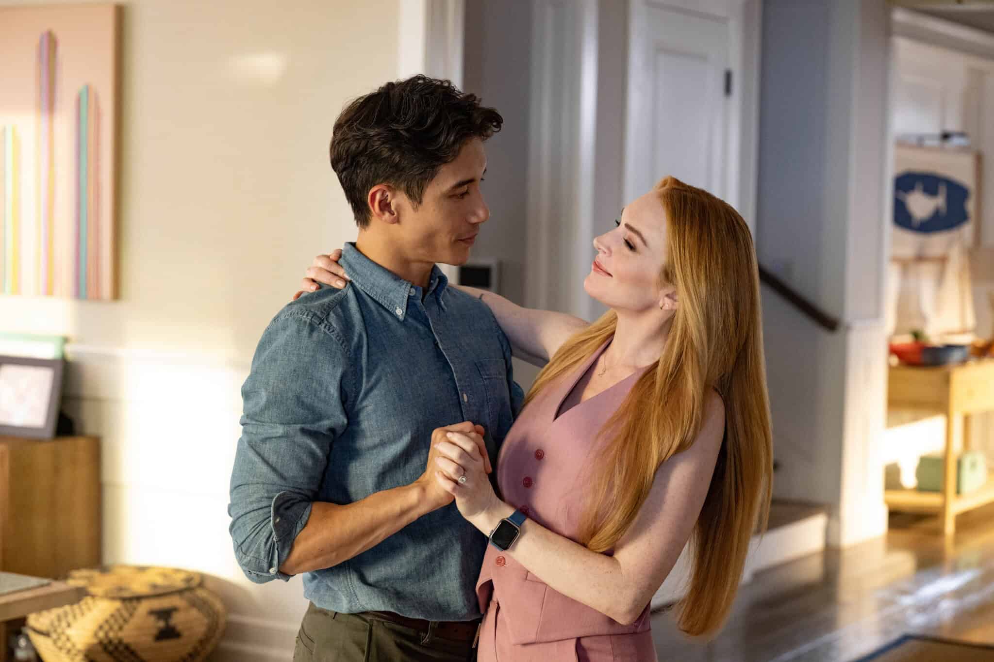 A young couple stands closely together in a warmly lit living room, holding hands and smiling while looking into each other’s eyes, appearing to dance or share an affectionate moment.