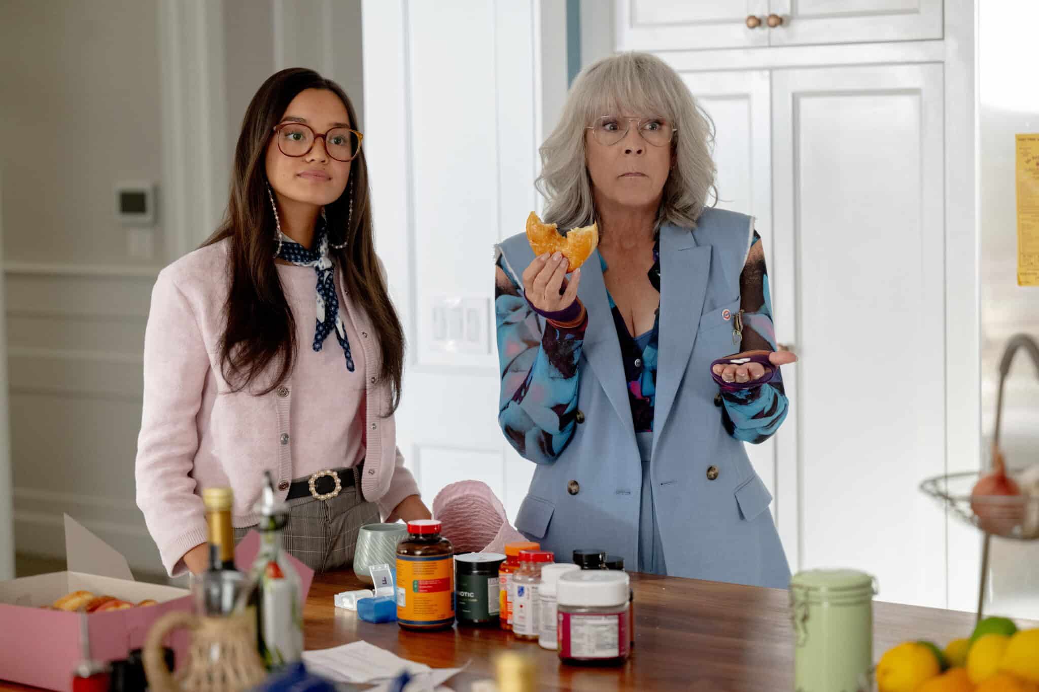 Two women stand in a kitchen. One wears glasses, a pink blazer, and patterned scarf, while the other has gray hair, glasses, a blue vest, and holds an orange slice. Various jars and bottles are on the counter in front of them.