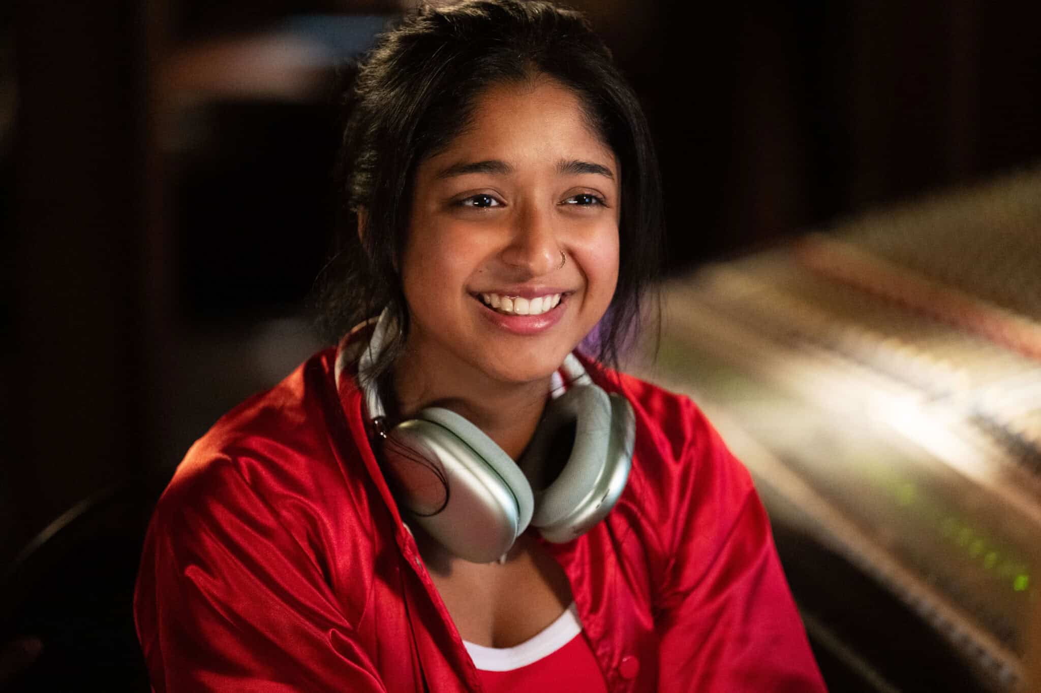 A young woman with long dark hair and a red jacket smiles while wearing large silver headphones around her neck, sitting in a dimly lit room with audio equipment in the background.