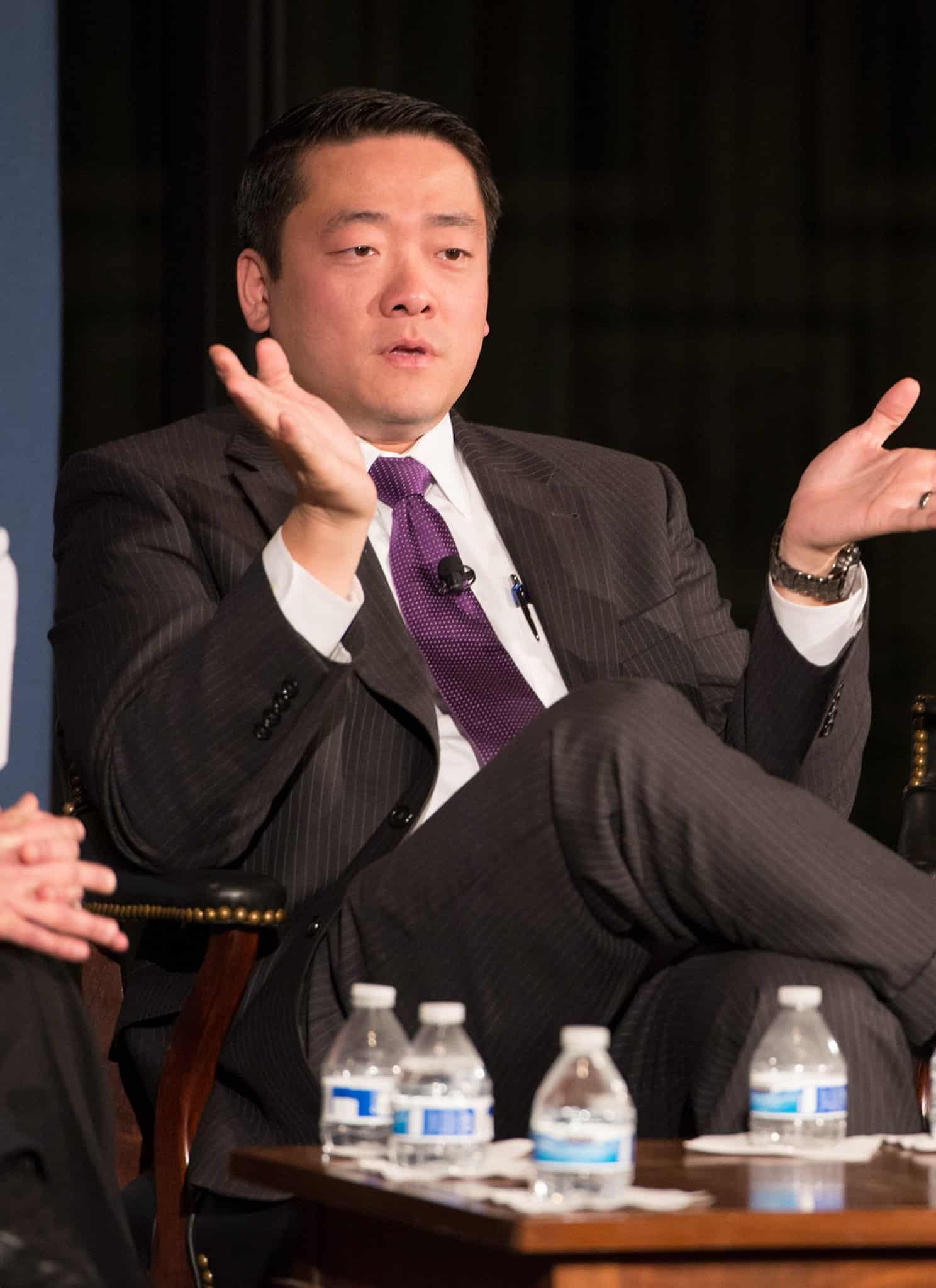 A man in a suit gestures with open hands while speaking, seated next to a woman in a black dress with a decorative neckline, during an event or panel discussion.