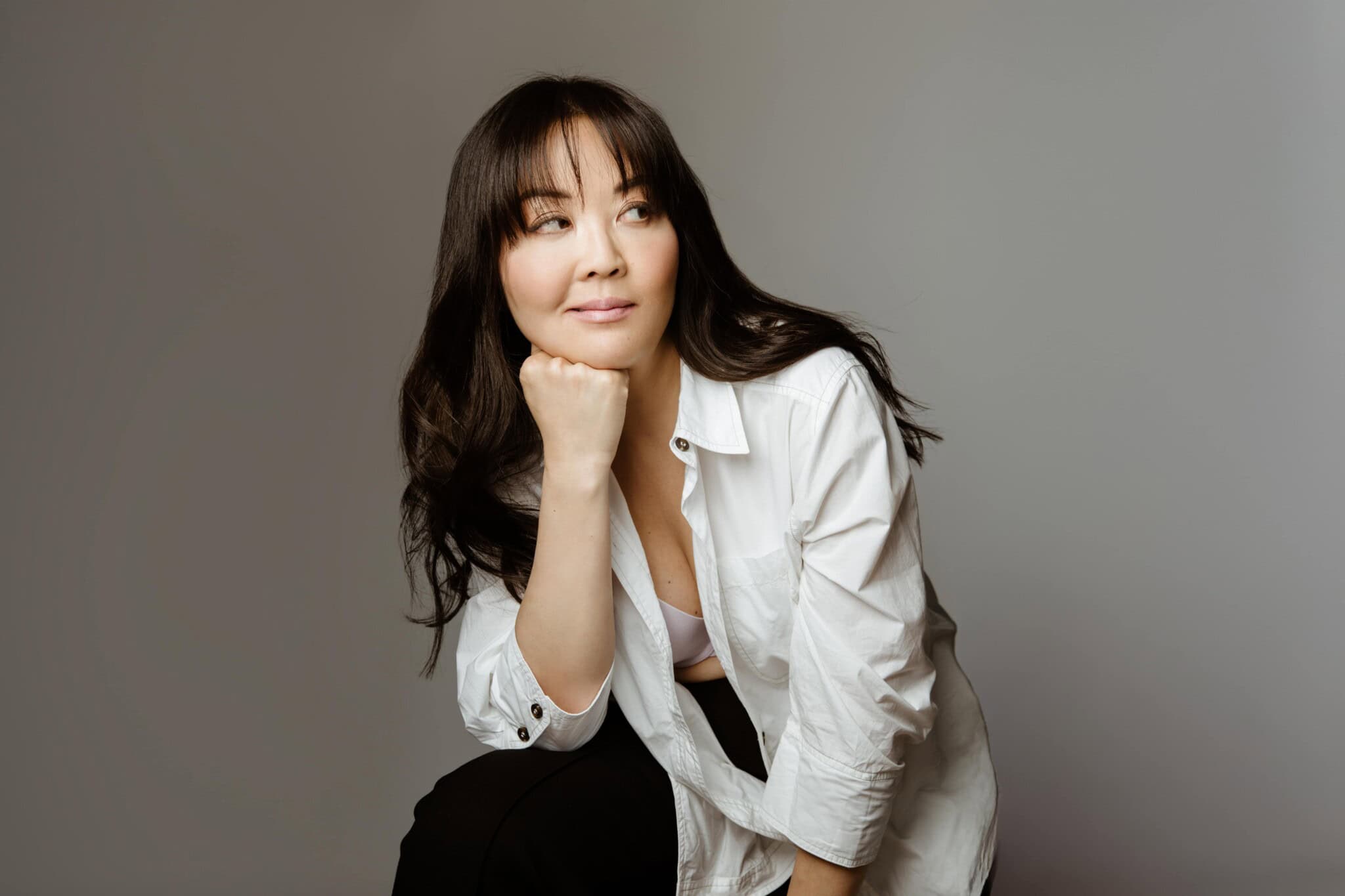 A woman with long dark hair wearing a white shirt leans forward, resting her chin on her hand, and looks thoughtfully to the side against a plain gray background.