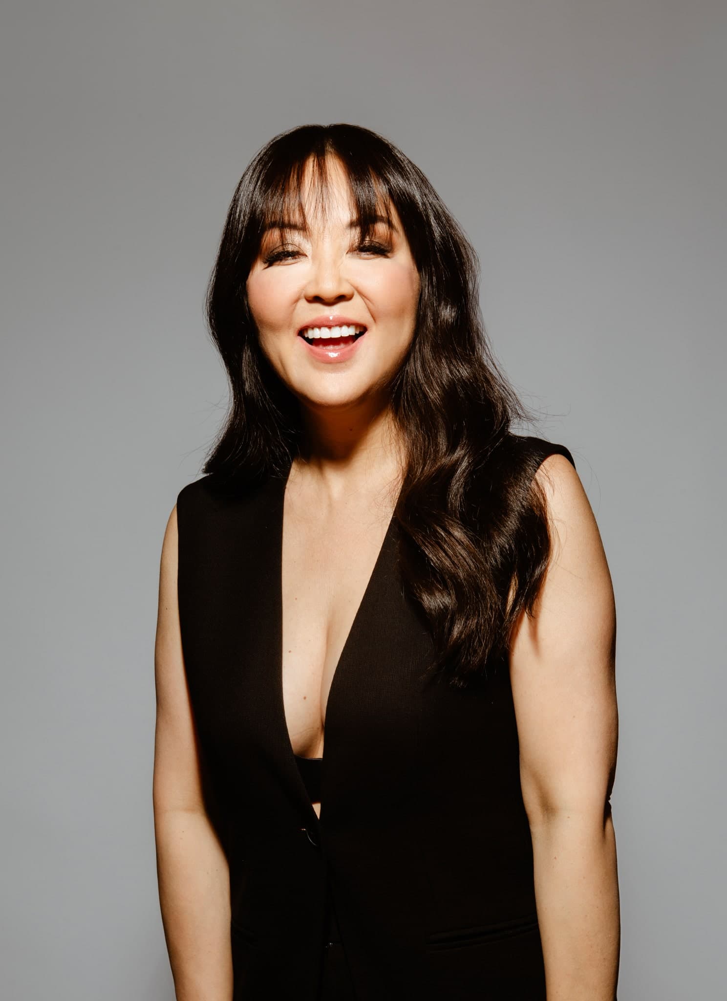 A woman with long, wavy dark hair and bangs smiles at the camera. She is wearing a sleeveless black outfit against a plain gray background.