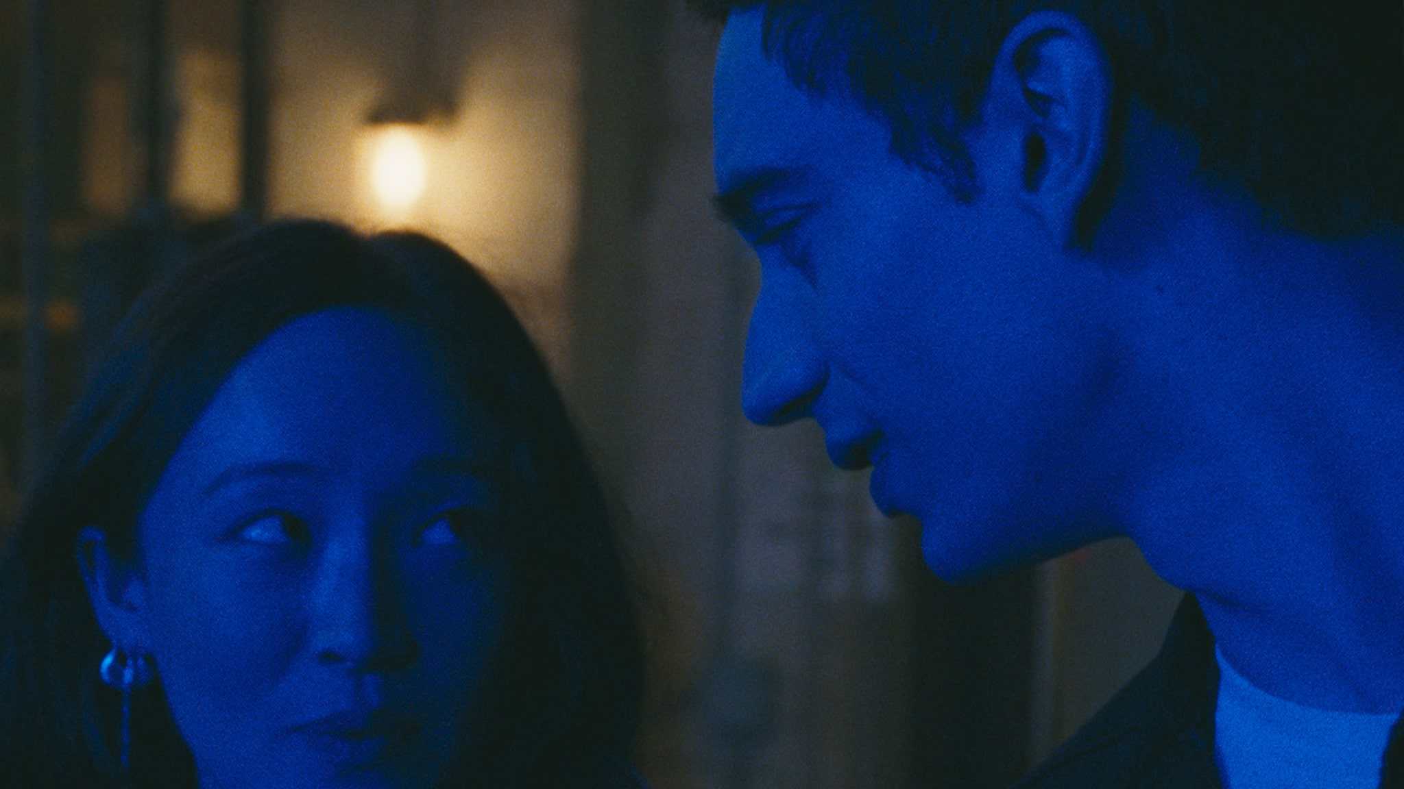 A woman and a man stand close together at night, illuminated by blue lighting. The woman looks at the man with a soft expression, while the man gazes intently at her. A blurred, warm light glows in the background.