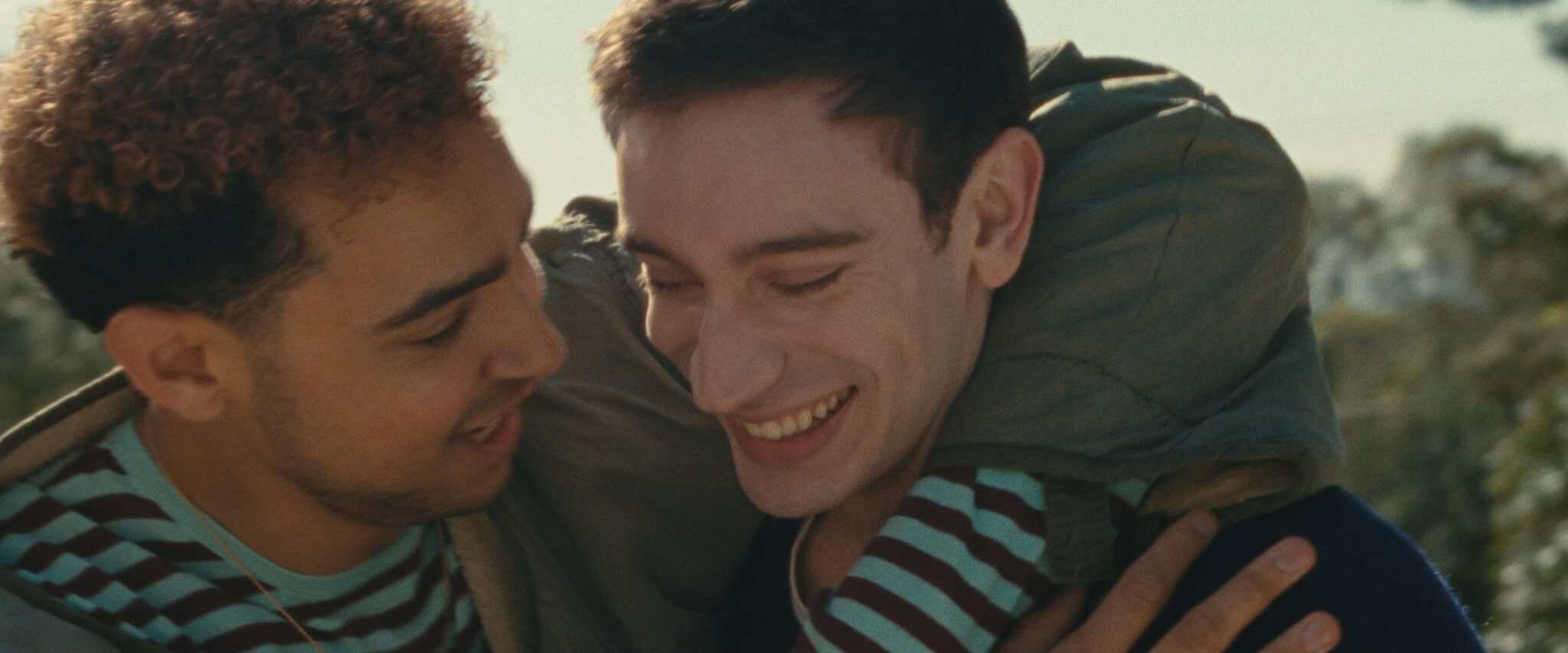 Two people smile and embrace outdoors. One person has their arm draped around the other, and both appear happy and connected. Trees and sunlight are visible in the background.
