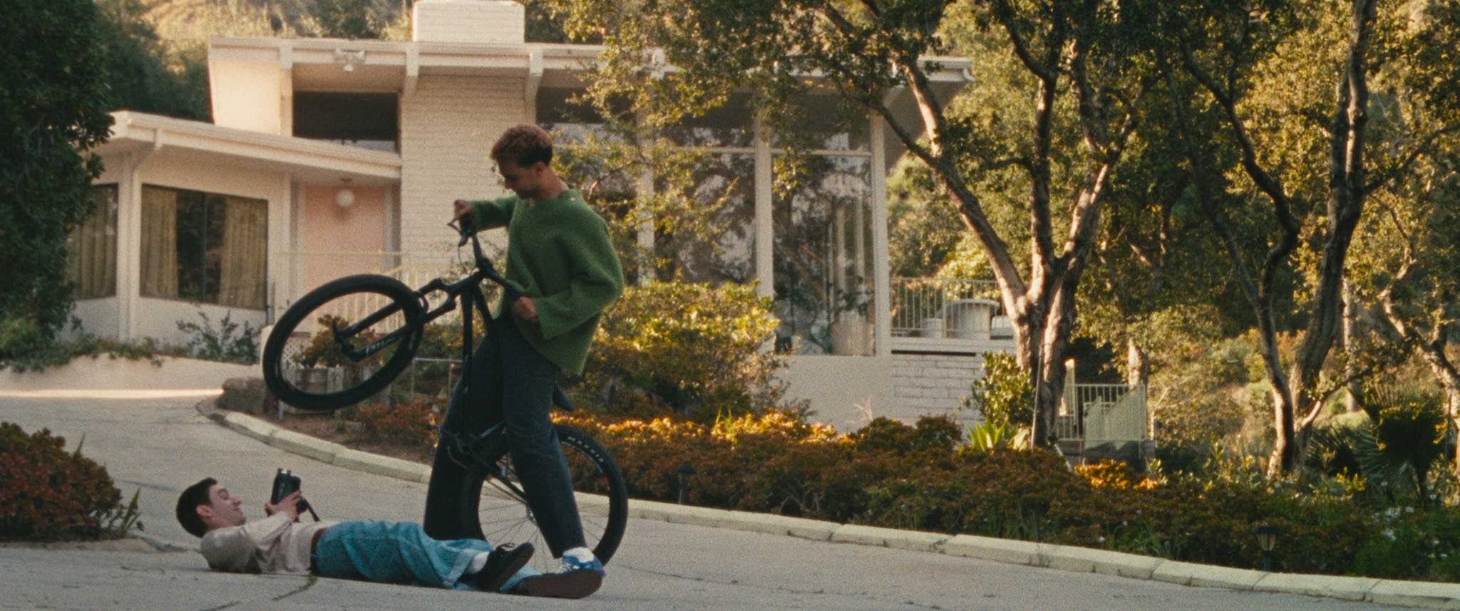 A person in a green sweater stands with a BMX bike, lifting its front wheel, while another person lies on their back on a driveway, holding a camera to take a photo. A modern house and trees are in the background.