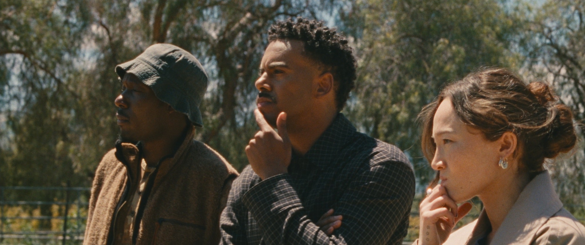 Three people stand outdoors, looking into the distance with thoughtful expressions. Two men, one wearing a bucket hat and the other in a checked shirt, and a woman with a beige coat are side by side with trees in the background.
